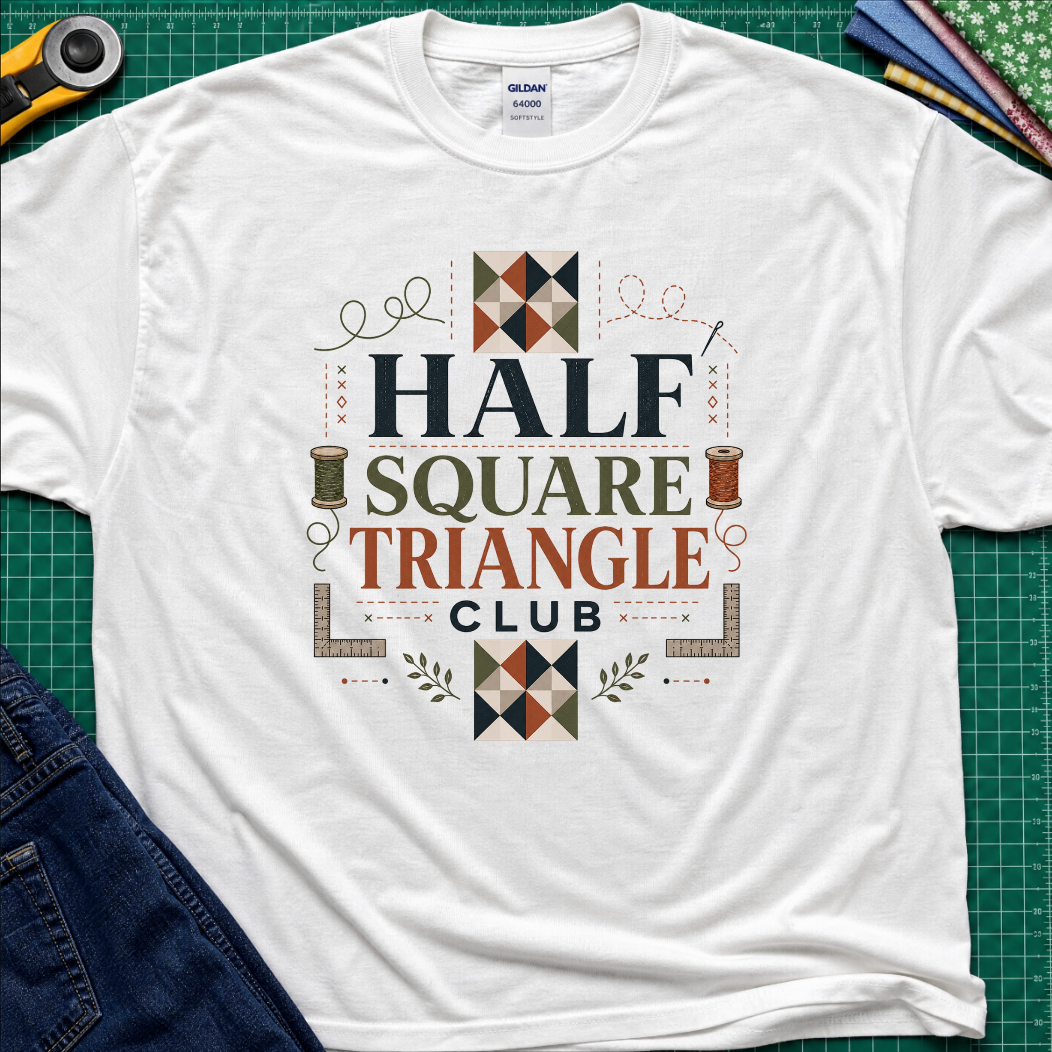 A white Gildan t-shirt with a 'Half Square Triangle Club' graphic, with quilt blocks, cotton spools and rulers. Laid flat on a green cutting mat with sewing props and jeans.