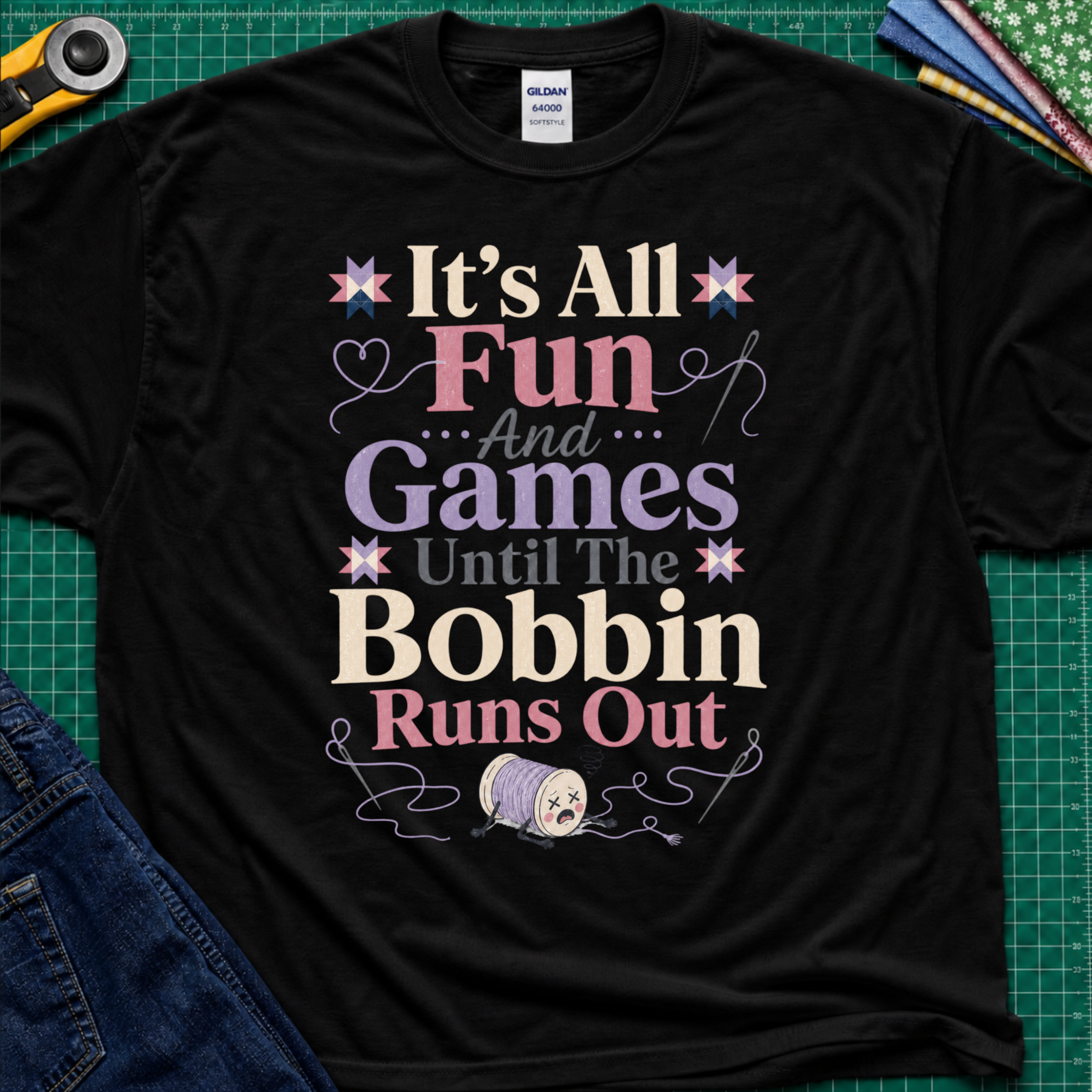 A black Gildan t-shirt with a colorful graphic print reading 'It's All Fun And Games Until The Bobbin Runs Out' in pink, purple, and cream lettering, styled flat on a green cutting mat with sewing tools and denim jeans.