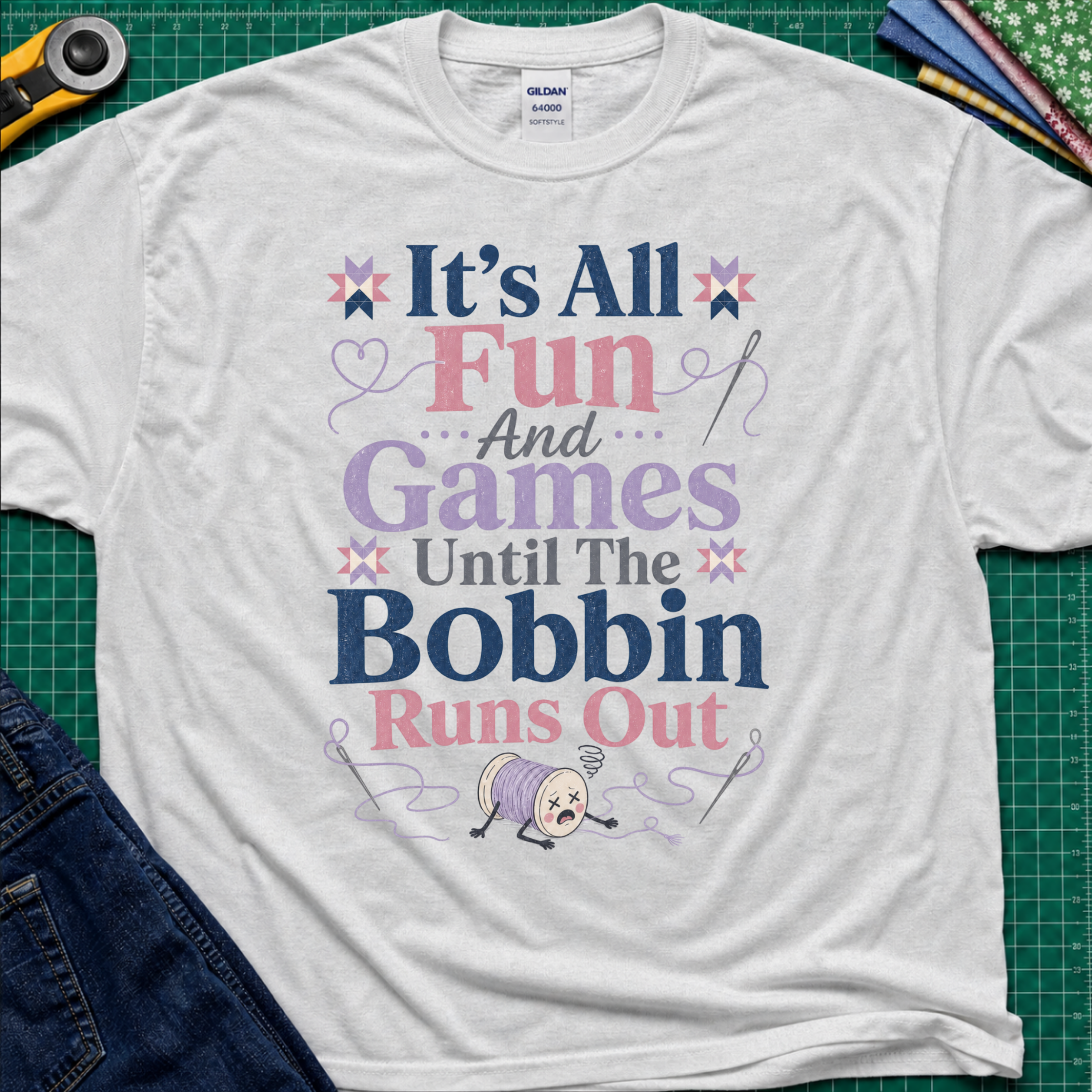 A light grey Gildan t-shirt with a colorful graphic print reading 'It's All Fun And Games Until The Bobbin Runs Out' in pink, purple, and navy lettering, styled flat on a green cutting mat with sewing tools and denim jeans.