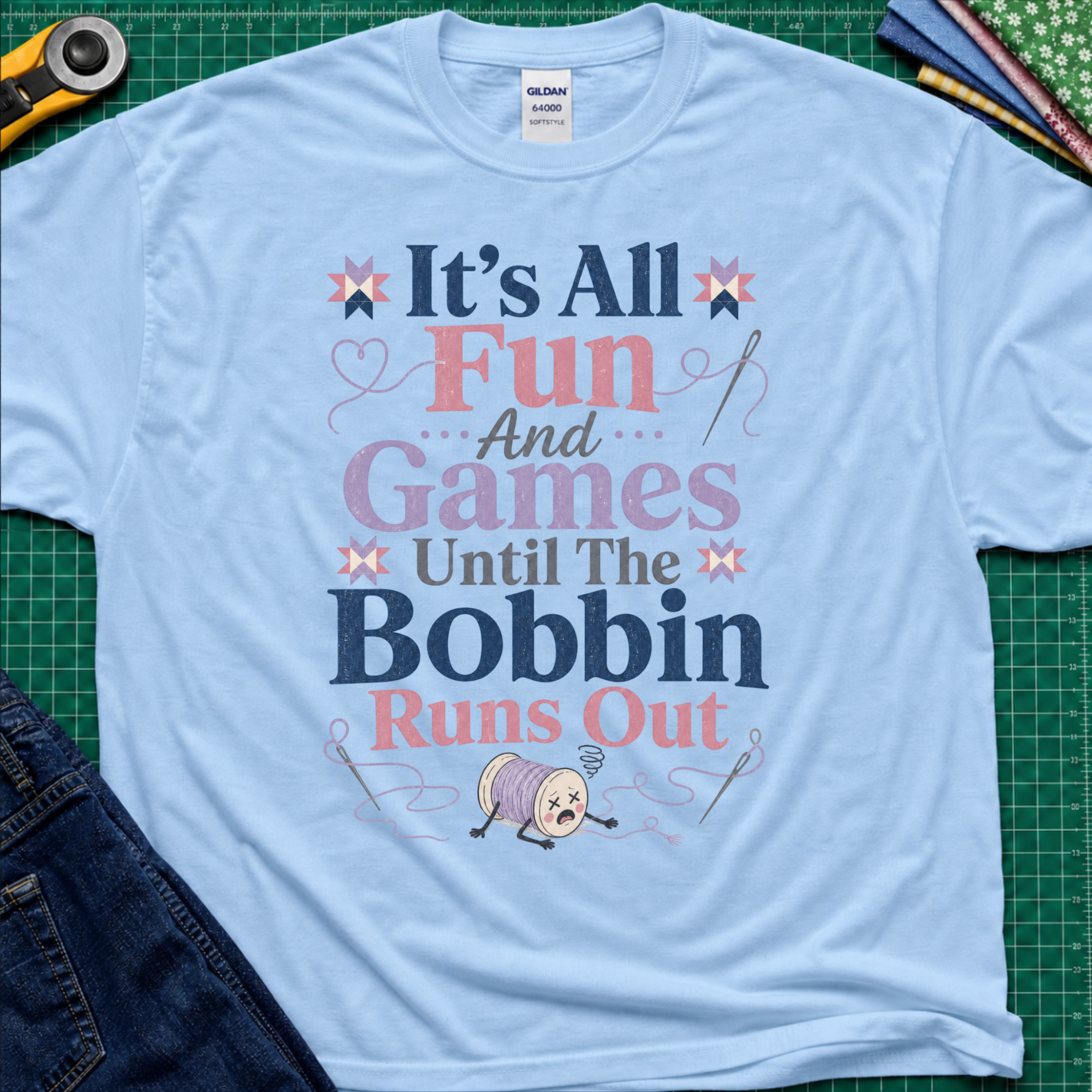 A light blue Gildan t-shirt with a colorful graphic print reading 'It's All Fun And Games Until The Bobbin Runs Out' in pink, purple, and navy lettering, styled flat on a green cutting mat with sewing tools and denim jeans.