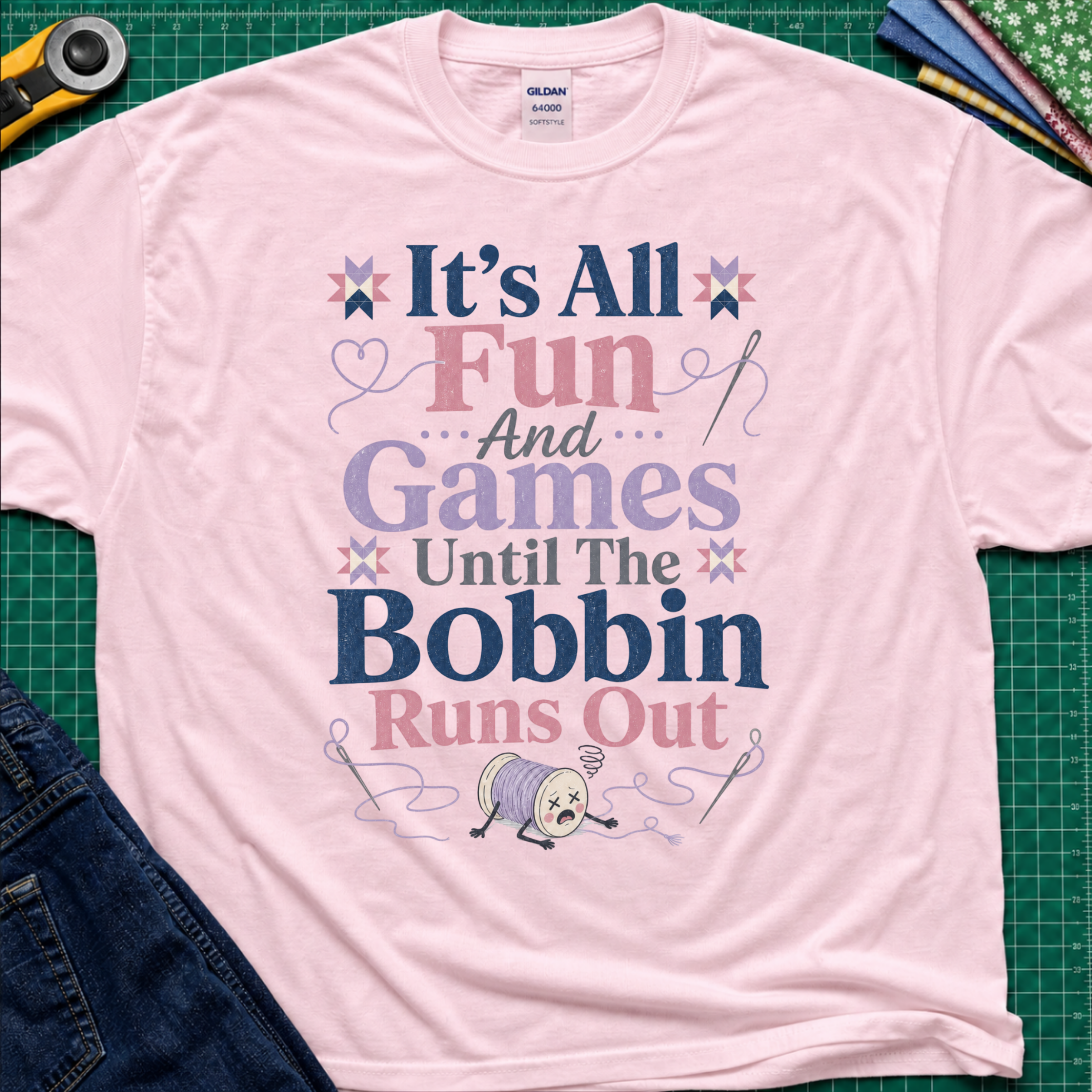 A light pink Gildan t-shirt with a colorful graphic print reading 'It's All Fun And Games Until The Bobbin Runs Out' in pink, purple, and navy lettering, styled flat on a green cutting mat with sewing tools and denim jeans.