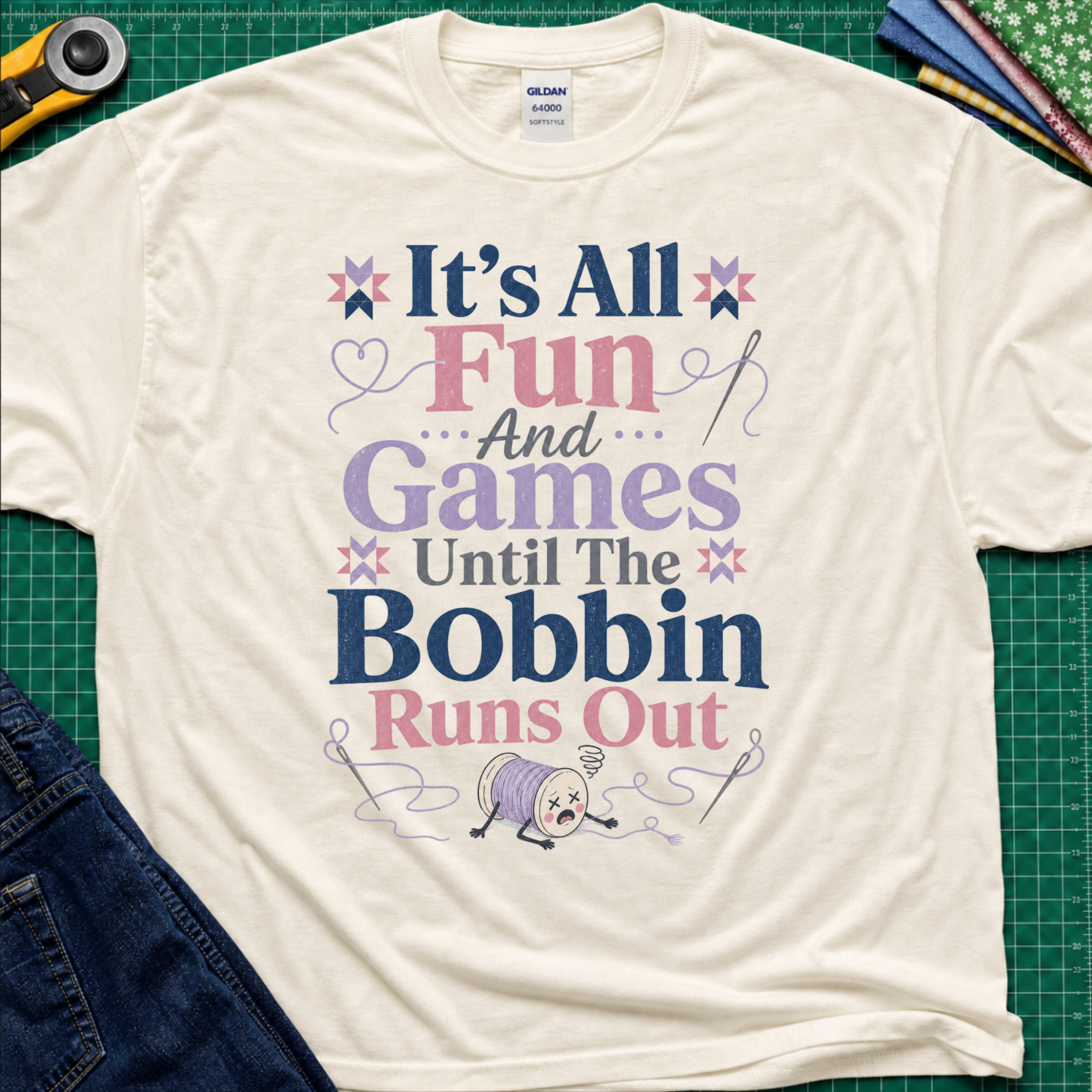A light cream Gildan t-shirt with a colorful graphic print reading 'It's All Fun And Games Until The Bobbin Runs Out' in pink, purple, and navy lettering, styled flat on a green cutting mat with sewing tools and denim jeans.