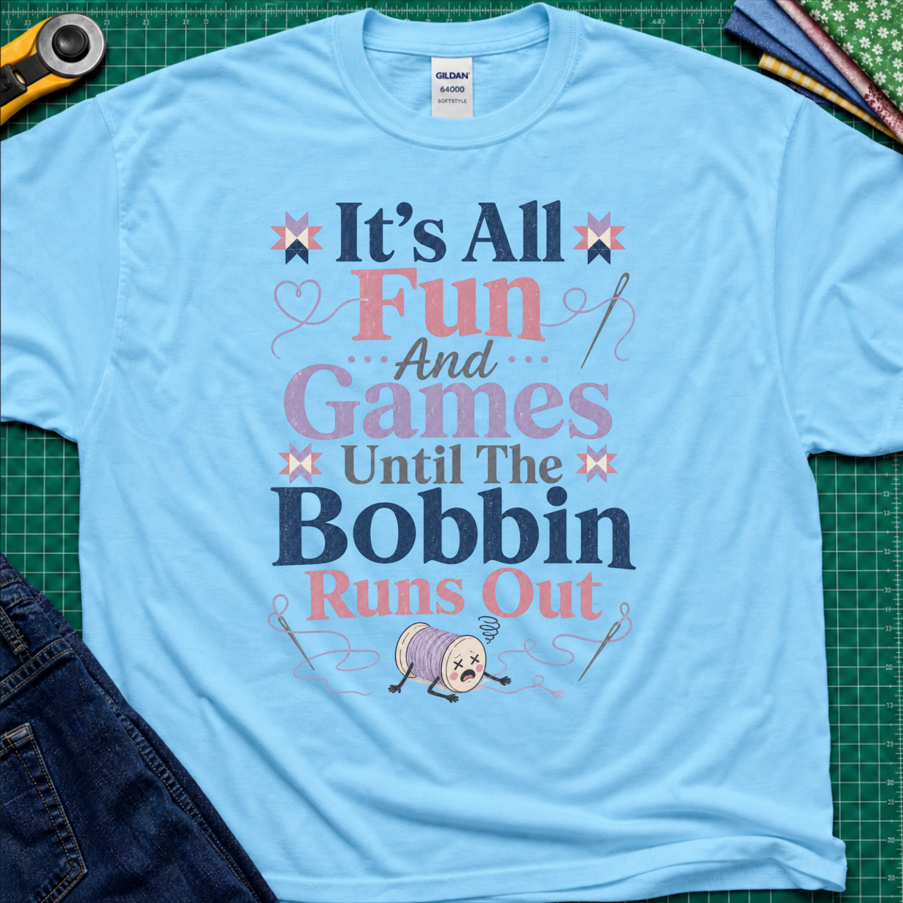 A sky blue Gildan t-shirt with a colorful graphic print reading 'It's All Fun And Games Until The Bobbin Runs Out' in pink, purple, and navy lettering, styled flat on a green cutting mat with sewing tools and denim jeans.