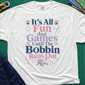 A white Gildan t-shirt with a colorful graphic print reading 'It's All Fun And Games Until The Bobbin Runs Out' in pink, purple, and navy lettering, styled flat on a green cutting mat with sewing tools and denim jeans.