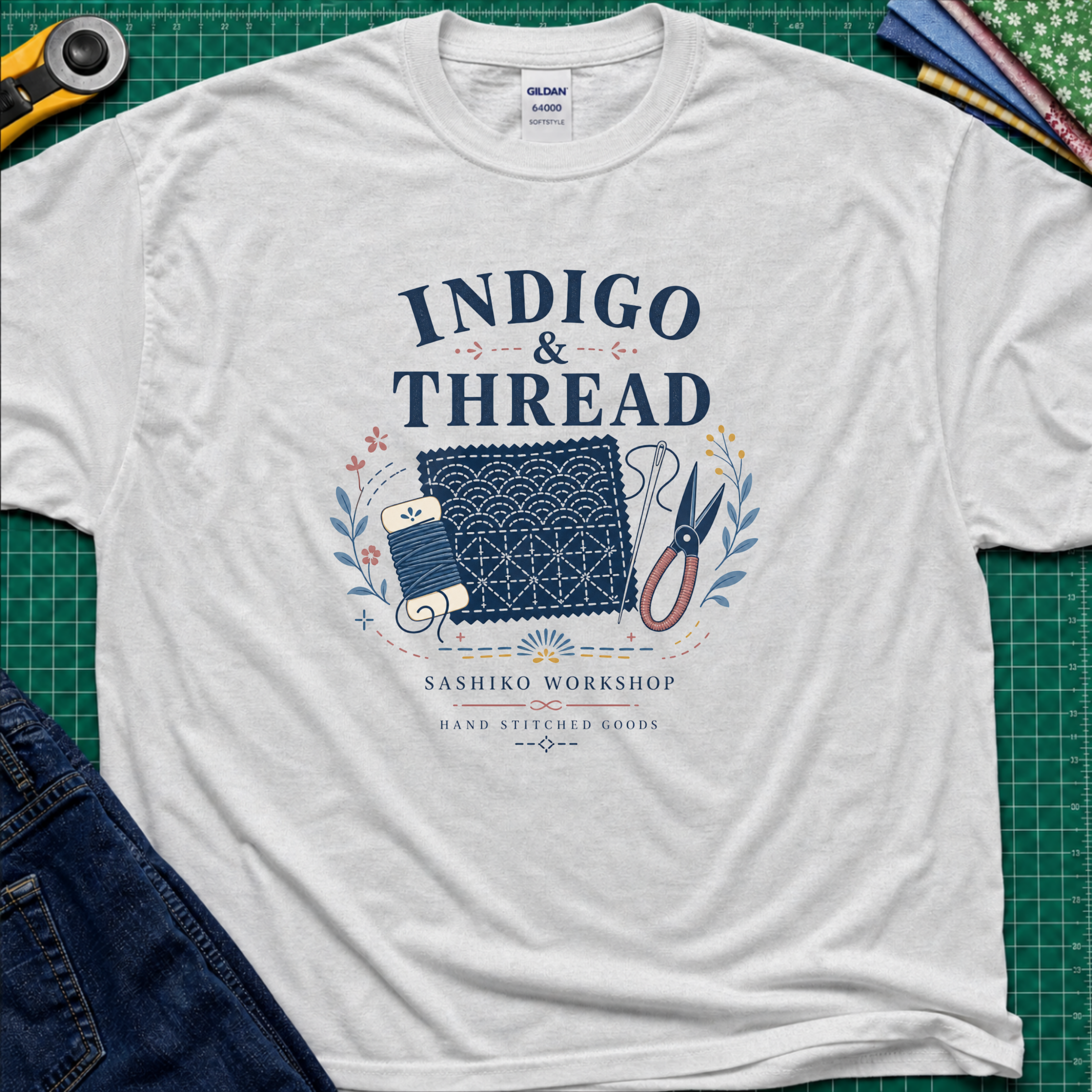 A light grey Gildan t-shirt that reads 'INDIGO & THREAD' with a sashiko stitched block, thread, scissors, needle and floral accents. The shirt lays flat on a green cutting mat with a rotary cutter, fabric stack and blue jeans partially visible in the corners.
