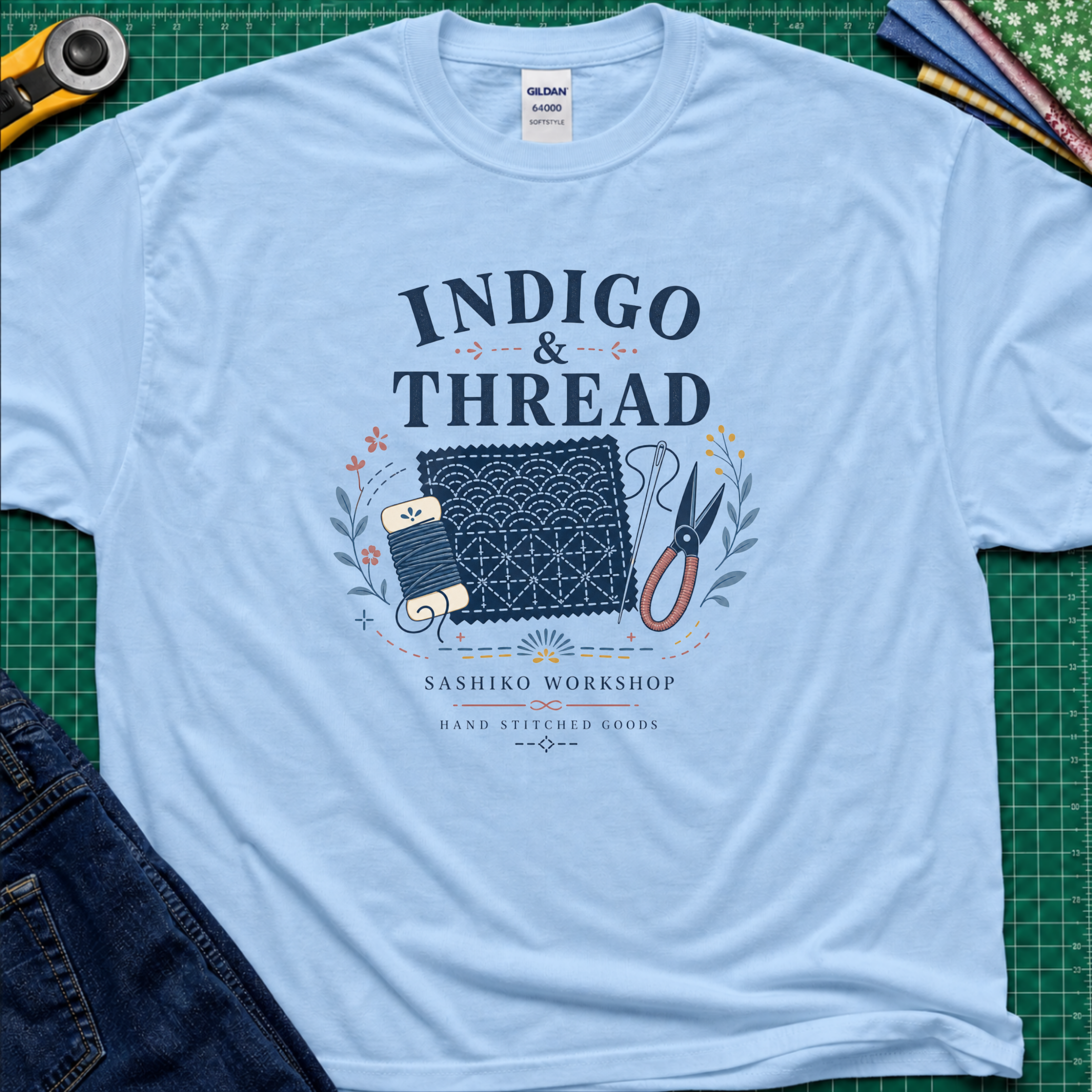 A light blue Gildan t-shirt that reads 'INDIGO & THREAD' with a sashiko stitched block, thread, scissors, needle and floral accents. The shirt lays flat on a green cutting mat with a rotary cutter, fabric stack and blue jeans partially visible in the corners.