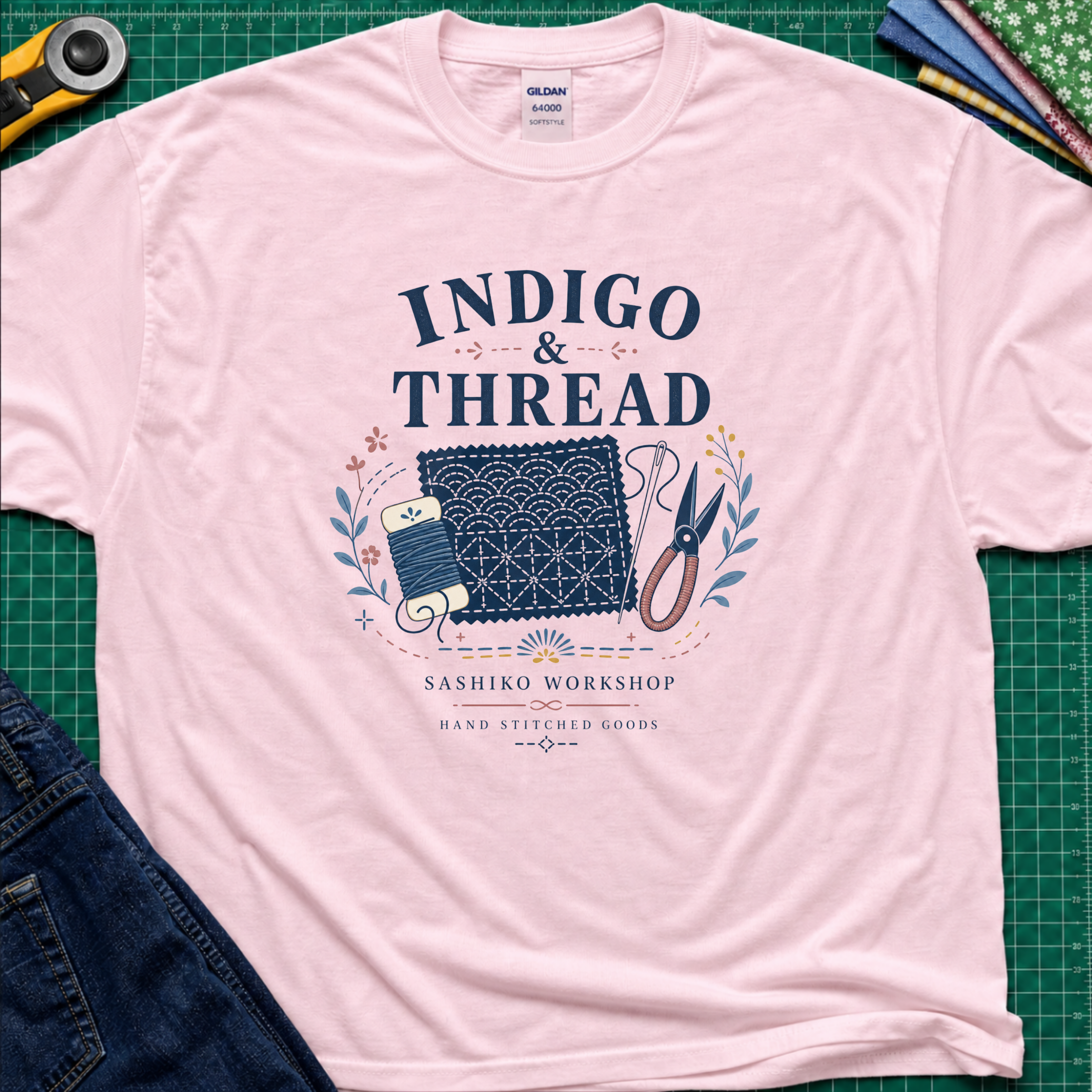 A light pink Gildan t-shirt that reads 'INDIGO & THREAD' with a sashiko stitched block, thread, scissors, needle and floral accents. The shirt lays flat on a green cutting mat with a rotary cutter, fabric stack and blue jeans partially visible in the corners.
