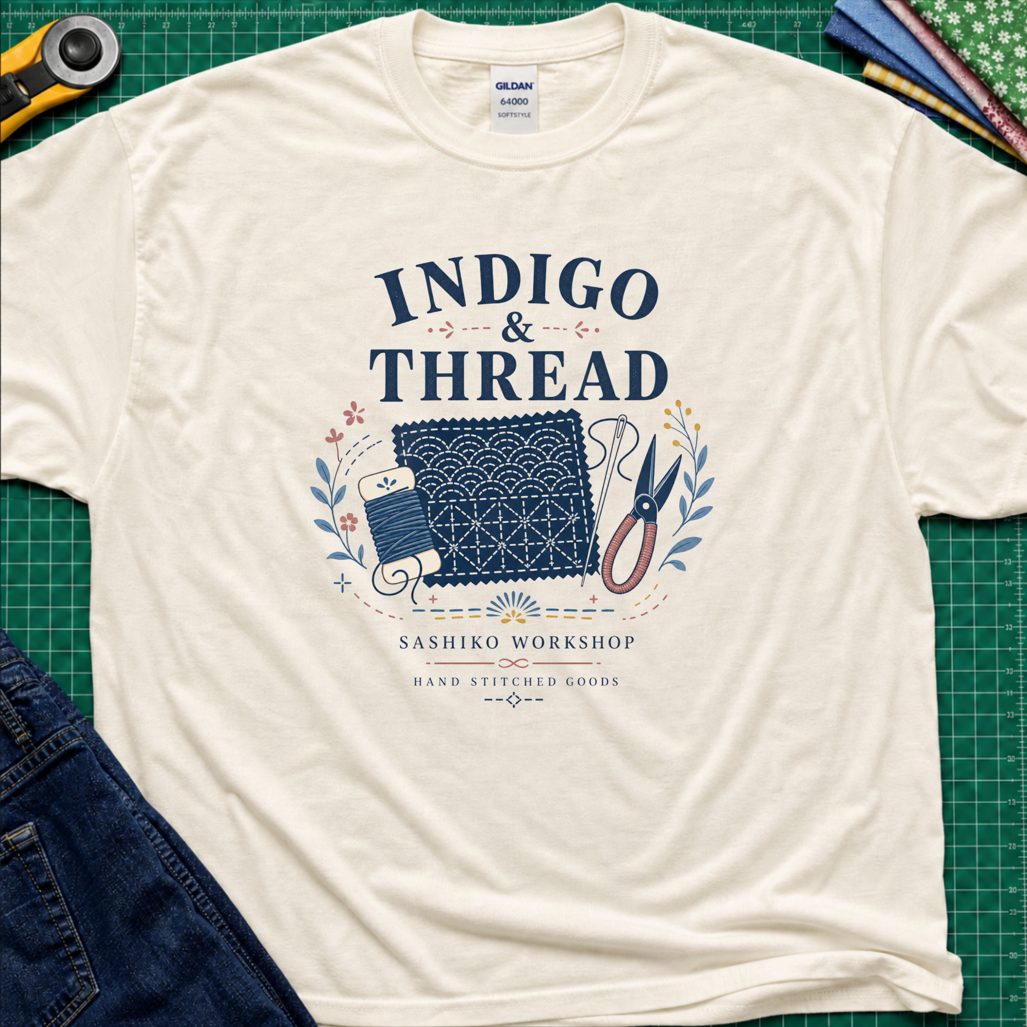 A light cream Gildan t-shirt that reads 'INDIGO & THREAD' with a sashiko stitched block, thread, scissors, needle and floral accents. The shirt lays flat on a green cutting mat with a rotary cutter, fabric stack and blue jeans partially visible in the corners.