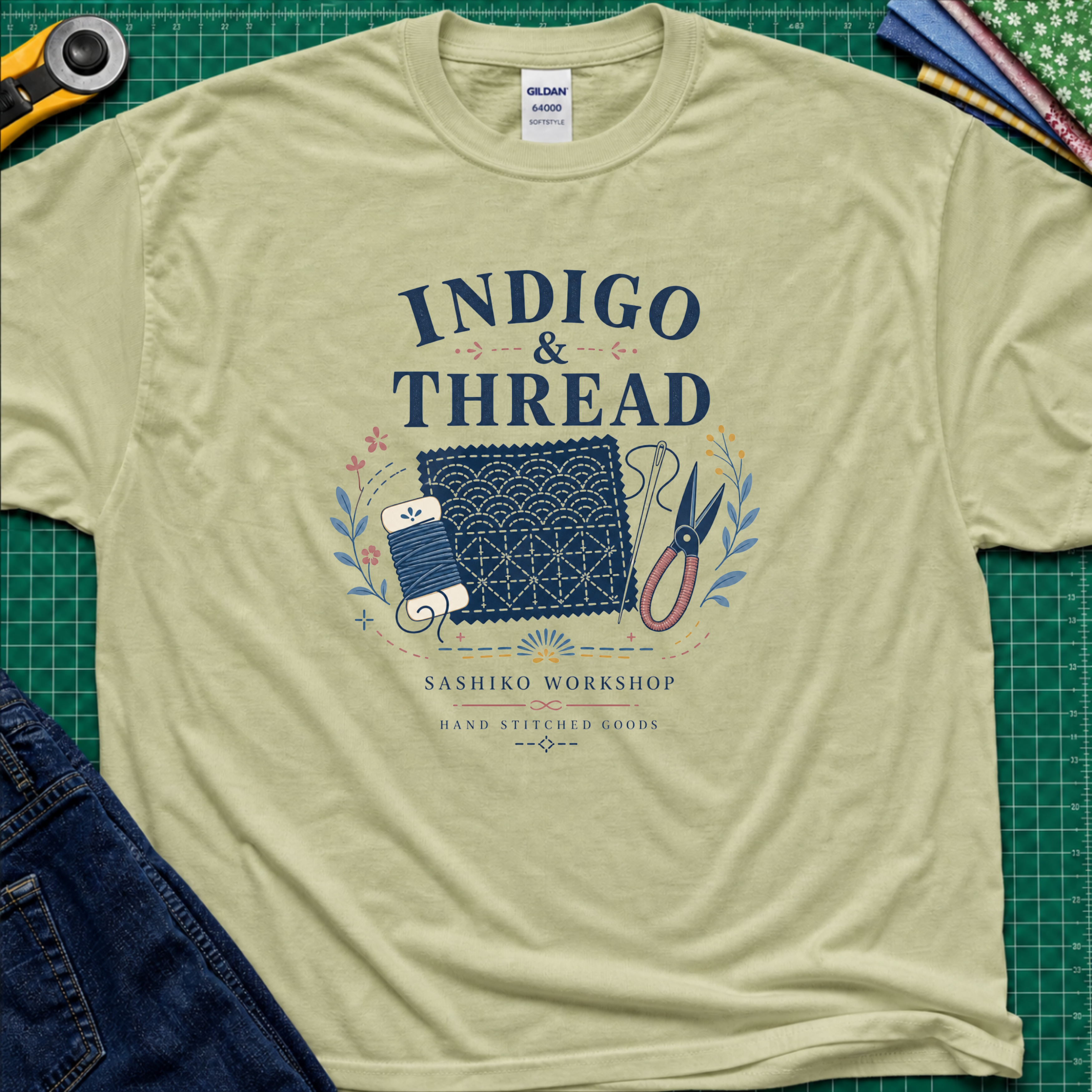 A light pistachio green Gildan t-shirt that reads 'INDIGO & THREAD' with a sashiko stitched block, thread, scissors, needle and floral accents. The shirt lays flat on a green cutting mat with a rotary cutter, fabric stack and blue jeans partially visible in the corners.