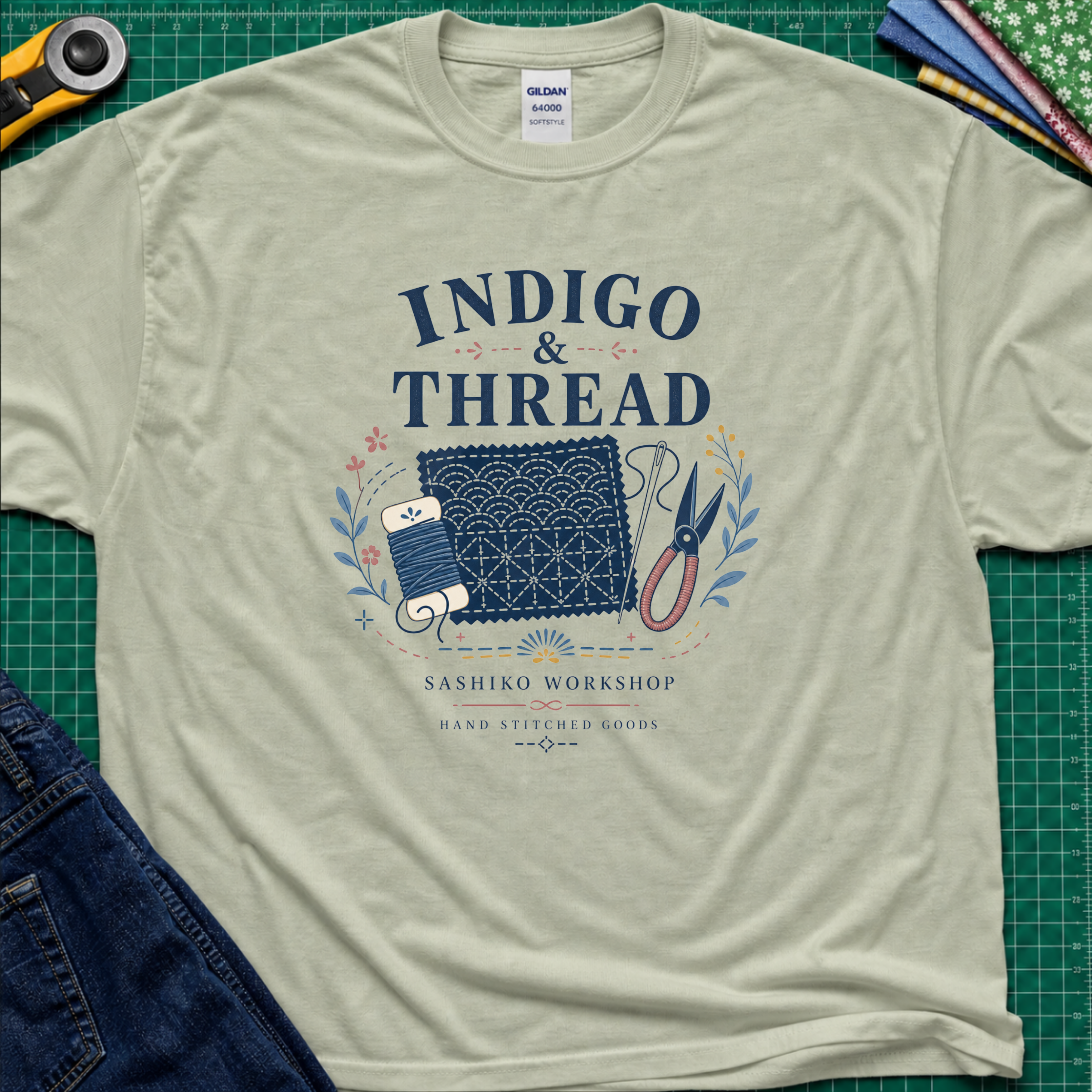 A light sage green Gildan t-shirt that reads 'INDIGO & THREAD' with a sashiko stitched block, thread, scissors, needle and floral accents. The shirt lays flat on a green cutting mat with a rotary cutter, fabric stack and blue jeans partially visible in the corners.