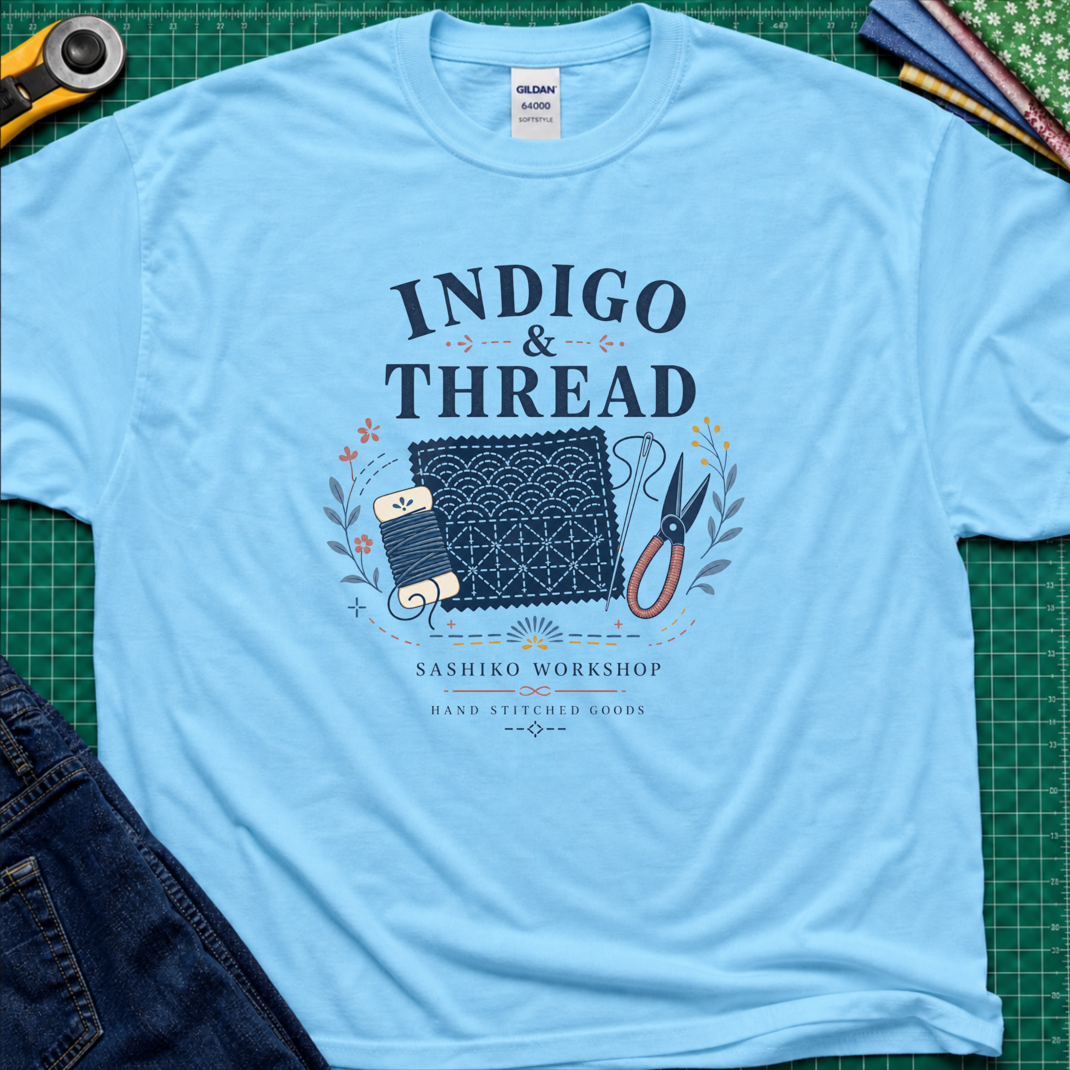 A sky blue Gildan t-shirt that reads 'INDIGO & THREAD' with a sashiko stitched block, thread, scissors, needle and floral accents. The shirt lays flat on a green cutting mat with a rotary cutter, fabric stack and blue jeans partially visible in the corners.