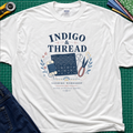 A white Gildan t-shirt that reads 'INDIGO & THREAD' with a sashiko stitched block, thread, scissors, needle and floral accents. The shirt lays flat on a green cutting mat with a rotary cutter, fabric stack and blue jeans partially visible in the corners.