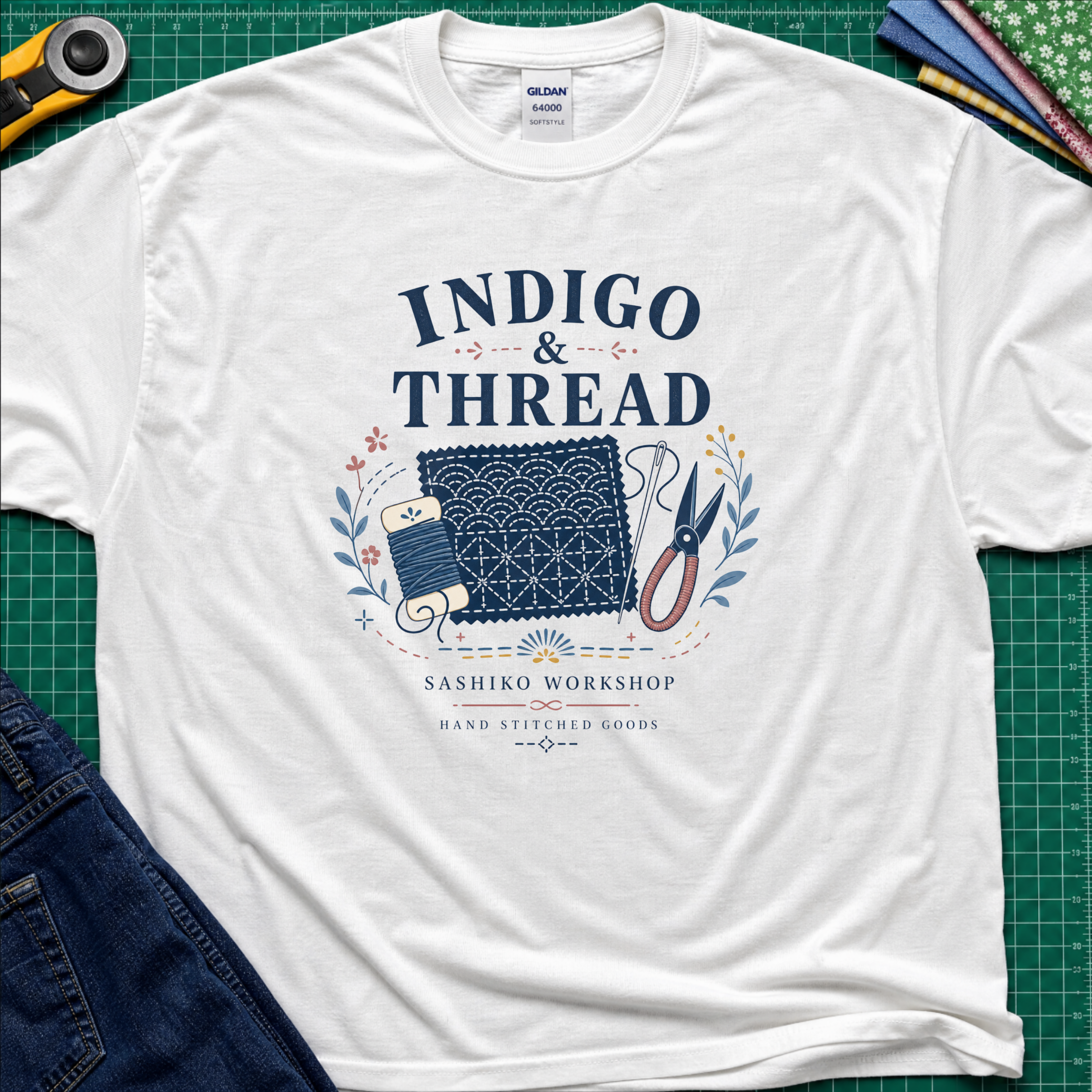 A white Gildan t-shirt that reads 'INDIGO & THREAD' with a sashiko stitched block, thread, scissors, needle and floral accents. The shirt lays flat on a green cutting mat with a rotary cutter, fabric stack and blue jeans partially visible in the corners.