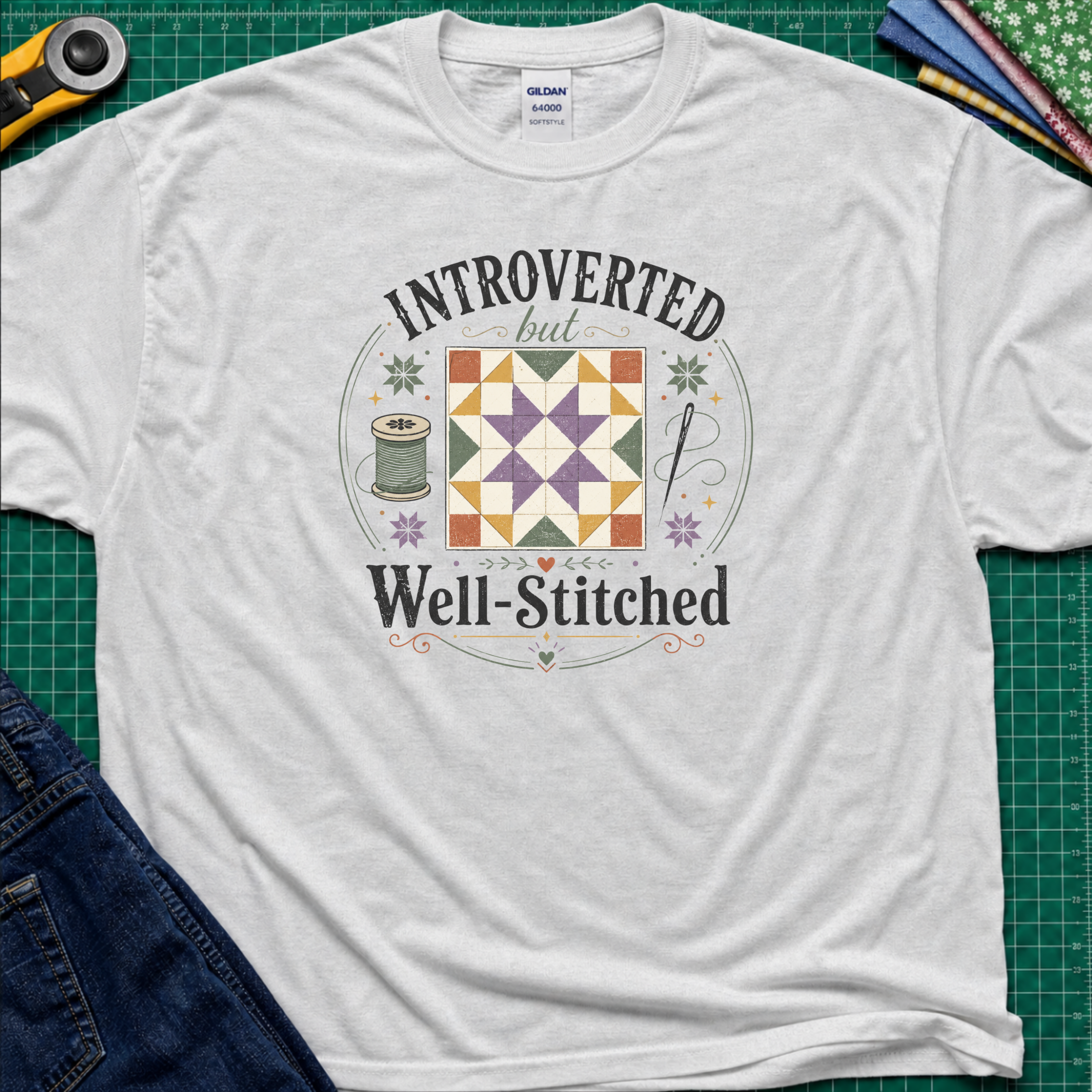 A light grey Gildan t-shirt laid flat on a green cutting mat, featuring a colorful retro graphic that reads 'INTROVERTED but Well-Stitched' around a quilt block. Sewing tools and blue jeans are partially visible as styled props.