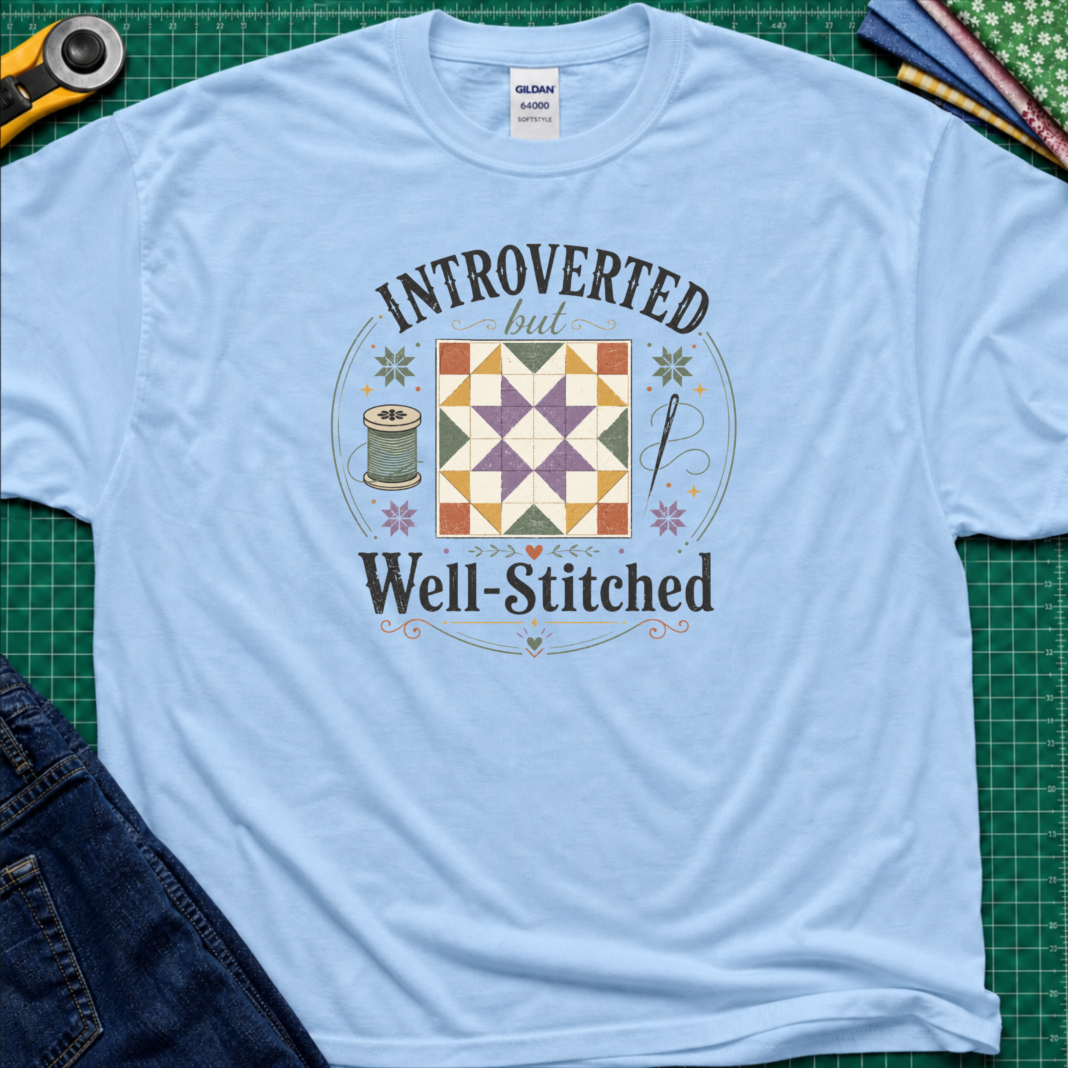 A light blue Gildan t-shirt laid flat on a green cutting mat, featuring a colorful retro graphic that reads 'INTROVERTED but Well-Stitched' around a quilt block. Sewing tools and blue jeans are partially visible as styled props.
