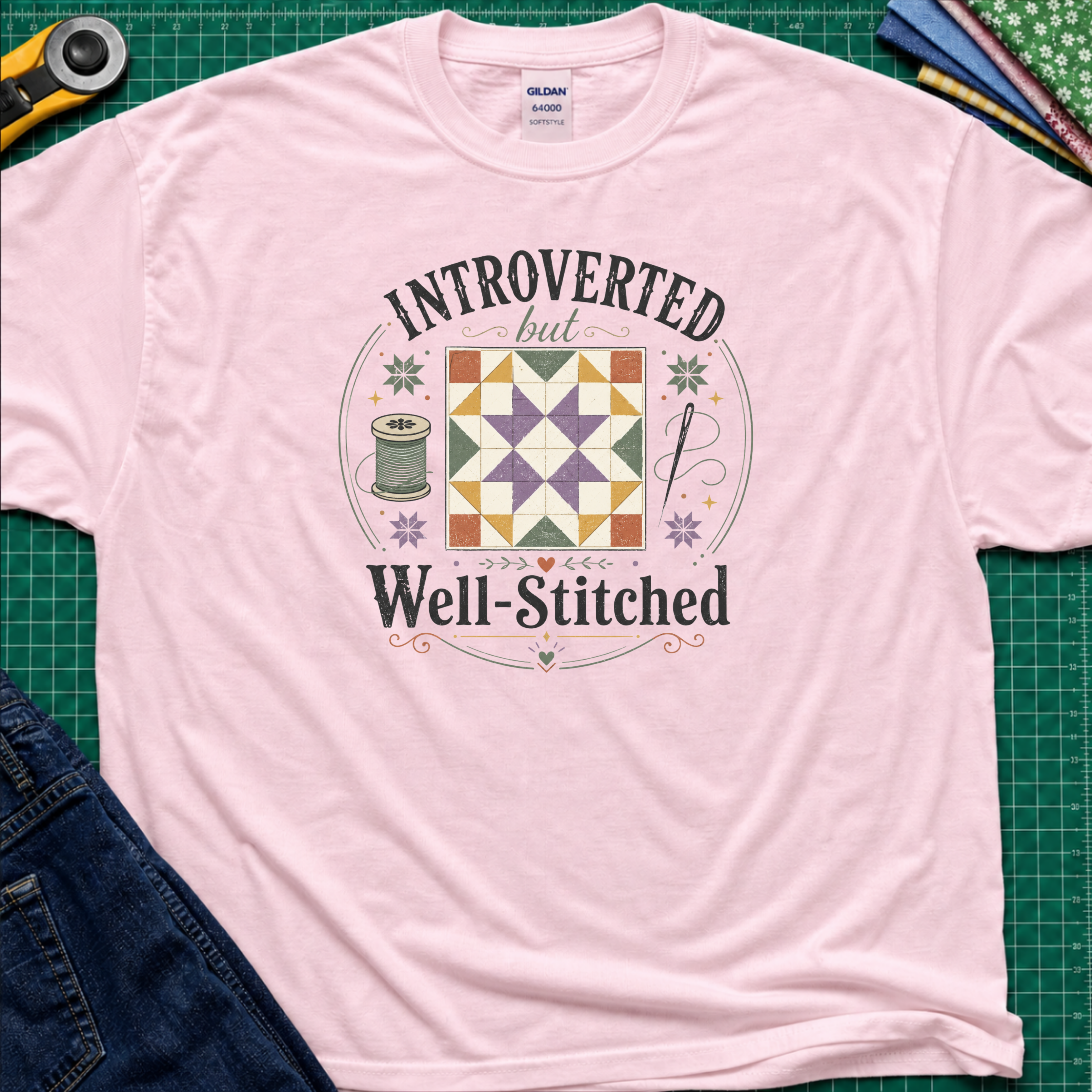 A light pink Gildan t-shirt laid flat on a green cutting mat, featuring a colorful retro graphic that reads 'INTROVERTED but Well-Stitched' around a quilt block. Sewing tools and blue jeans are partially visible as styled props.