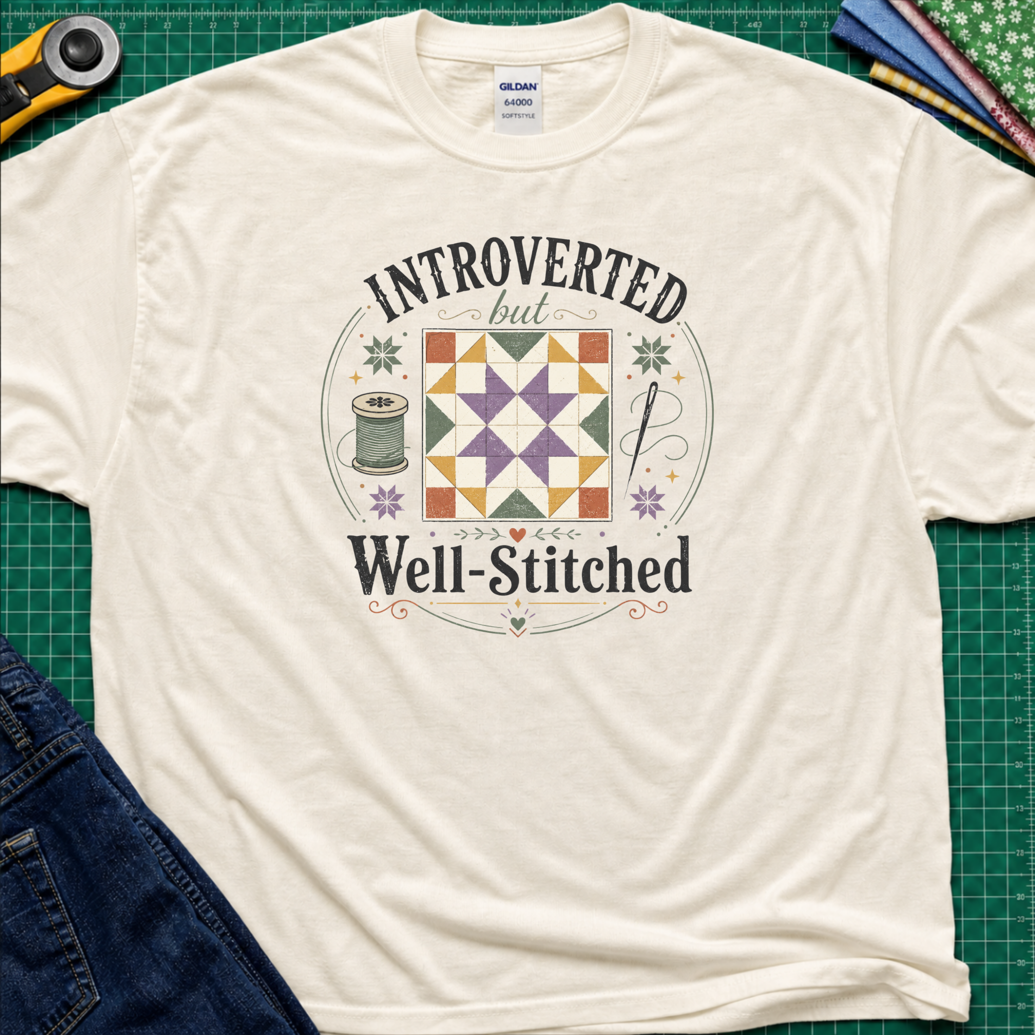 A light cream Gildan t-shirt laid flat on a green cutting mat, featuring a colorful retro graphic that reads 'INTROVERTED but Well-Stitched' around a quilt block. Sewing tools and blue jeans are partially visible as styled props.