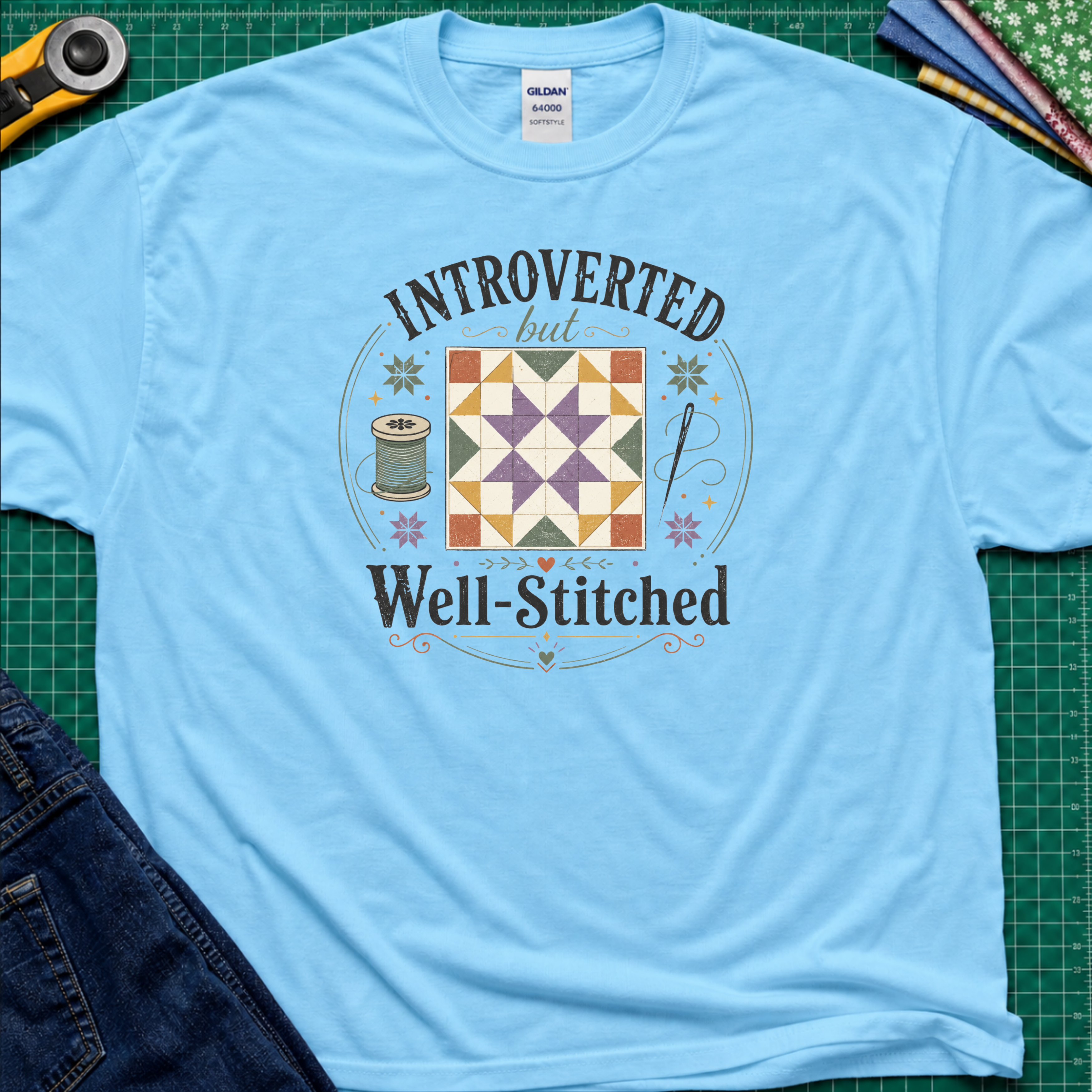 A sky blue Gildan t-shirt laid flat on a green cutting mat, featuring a colorful retro graphic that reads 'INTROVERTED but Well-Stitched' around a quilt block. Sewing tools and blue jeans are partially visible as styled props.