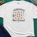 A white Gildan t-shirt laid flat on a green cutting mat, featuring a colorful retro graphic that reads 'INTROVERTED but Well-Stitched' around a quilt block. Sewing tools and blue jeans are partially visible as styled props.