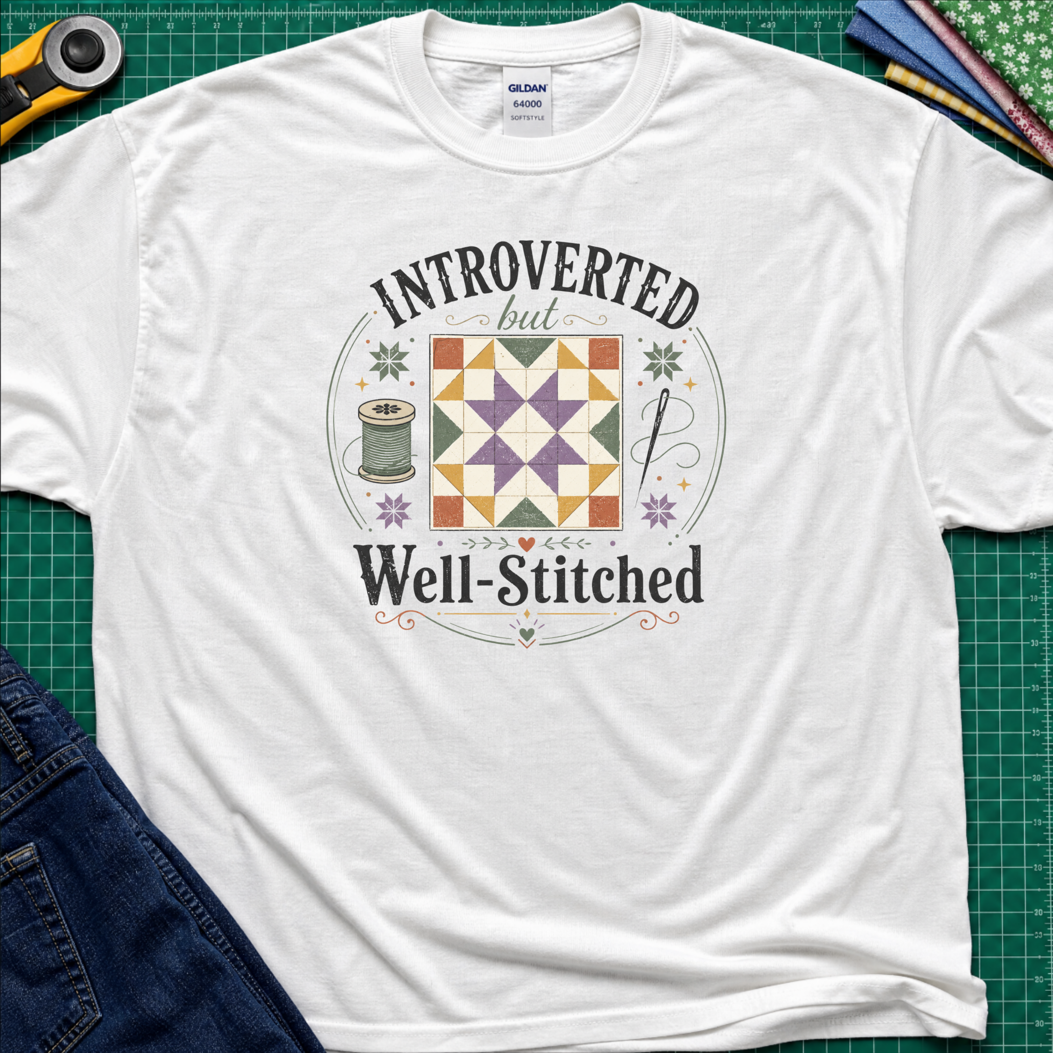 A white Gildan t-shirt laid flat on a green cutting mat, featuring a colorful retro graphic that reads 'INTROVERTED but Well-Stitched' around a quilt block. Sewing tools and blue jeans are partially visible as styled props.