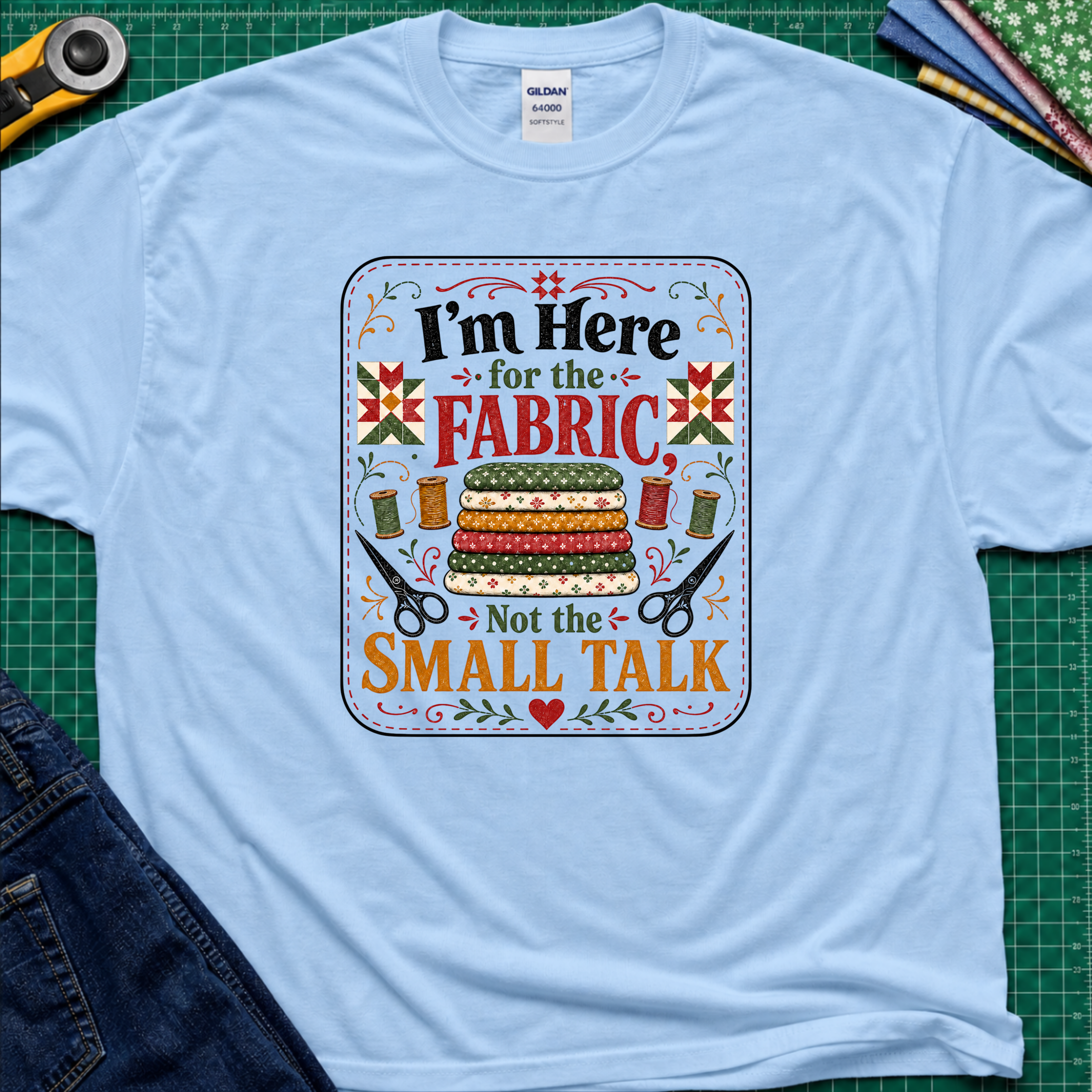A light blue Gildan t-shirt featuring a colorful graphic print with the text 'I'm Here for the FABRIC Not the SMALL TALK' and an illustration of stacked fabric bolts, scissors, and thread spools. The shirt is laid flat on a green cutting mat with a rotary cutter, fabric stack and blue jeans partially visible in the corners.