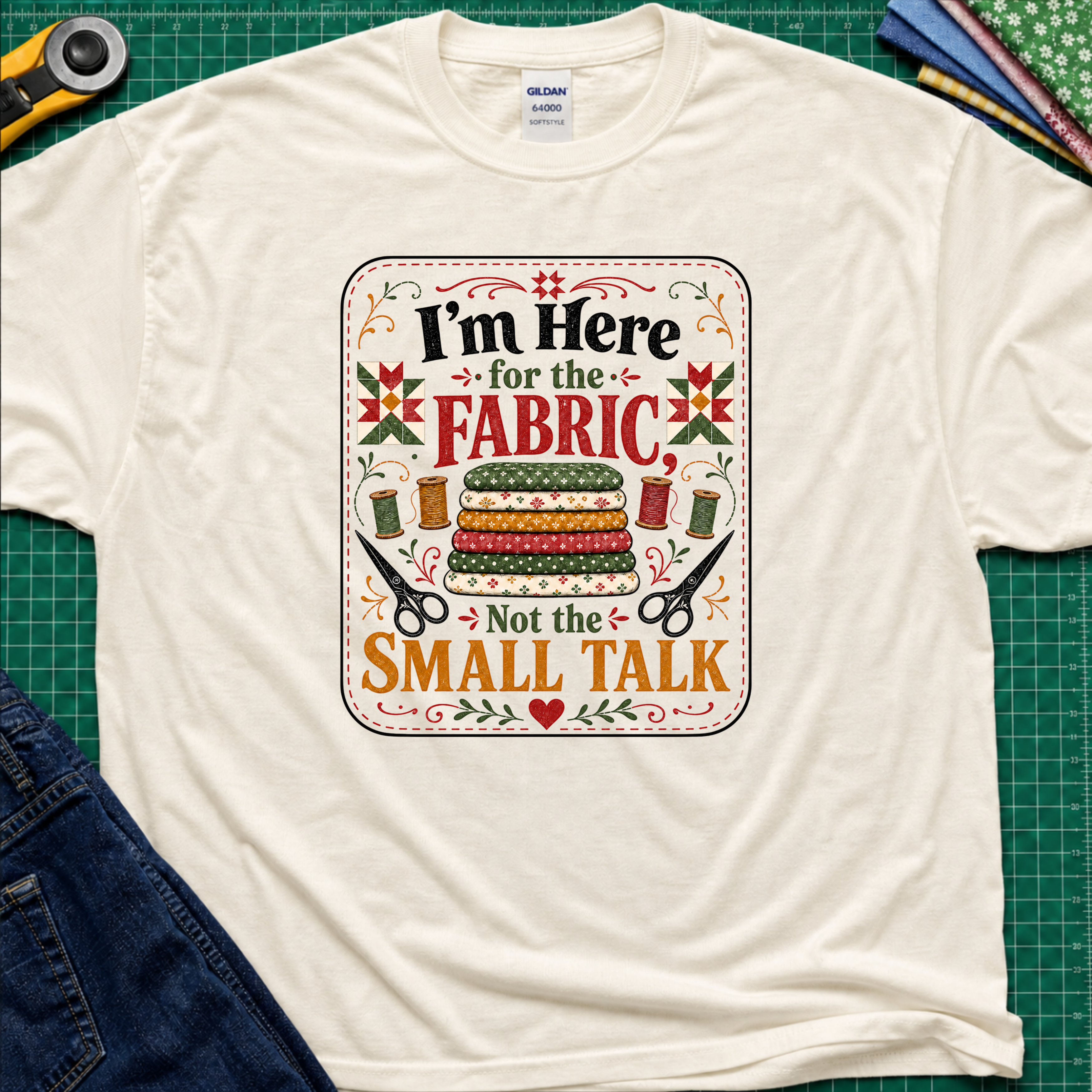 A light cream Gildan t-shirt featuring a colorful graphic print with the text 'I'm Here for the FABRIC Not the SMALL TALK' and an illustration of stacked fabric bolts, scissors, and thread spools. The shirt is laid flat on a green cutting mat with a rotary cutter, fabric stack and blue jeans partially visible in the corners.