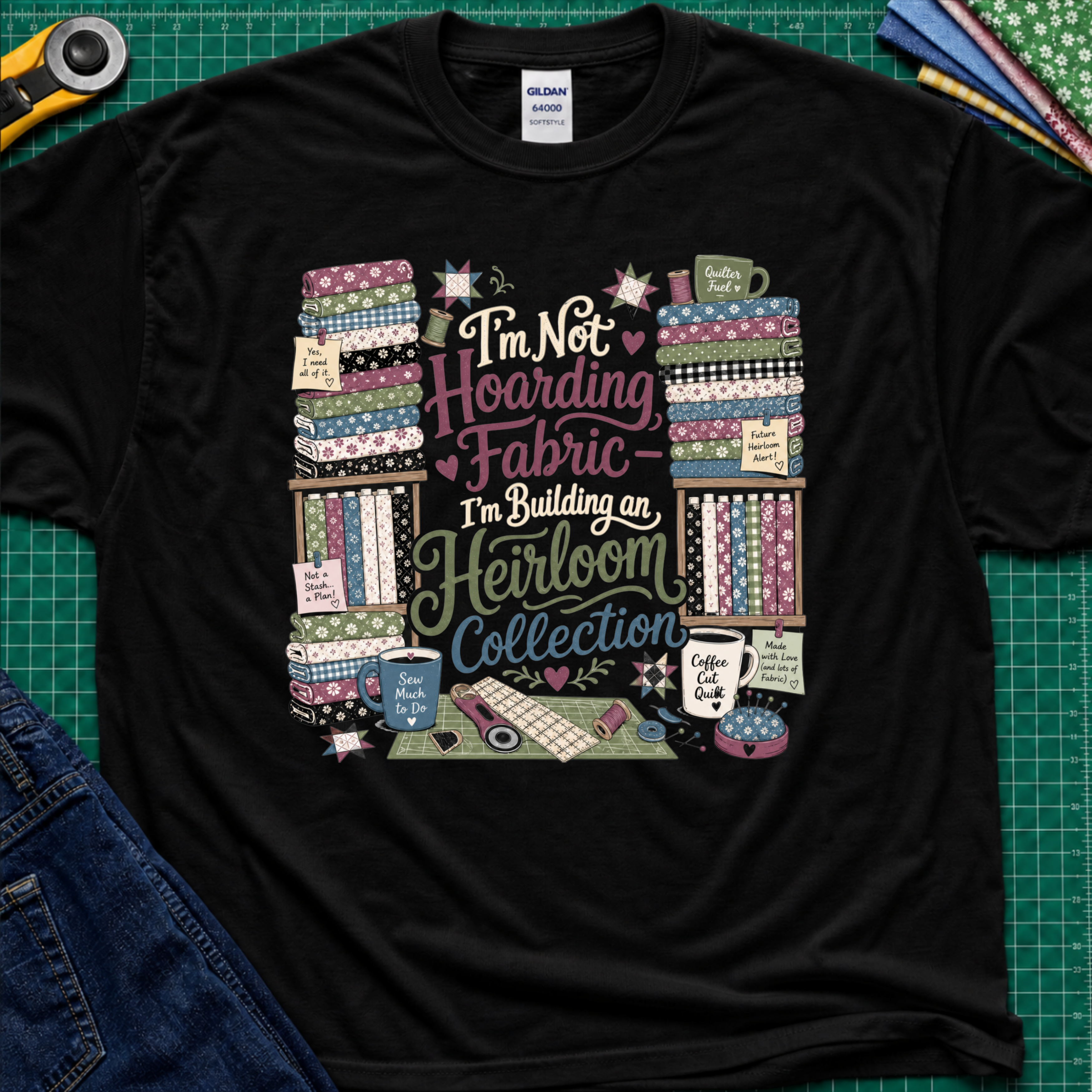 A black Gildan t-shirt  featuring a colorful print with fabric stacks, coffee cups, stars and quilting tools with text that reads, 'I'm Not Hoarding Fabric - I'm Building an Heirloom Collection'. The t-shirt lays flat on a green cutting mat with a rotary cutter, folded fabric and blue jeans partially visible in the corners.
