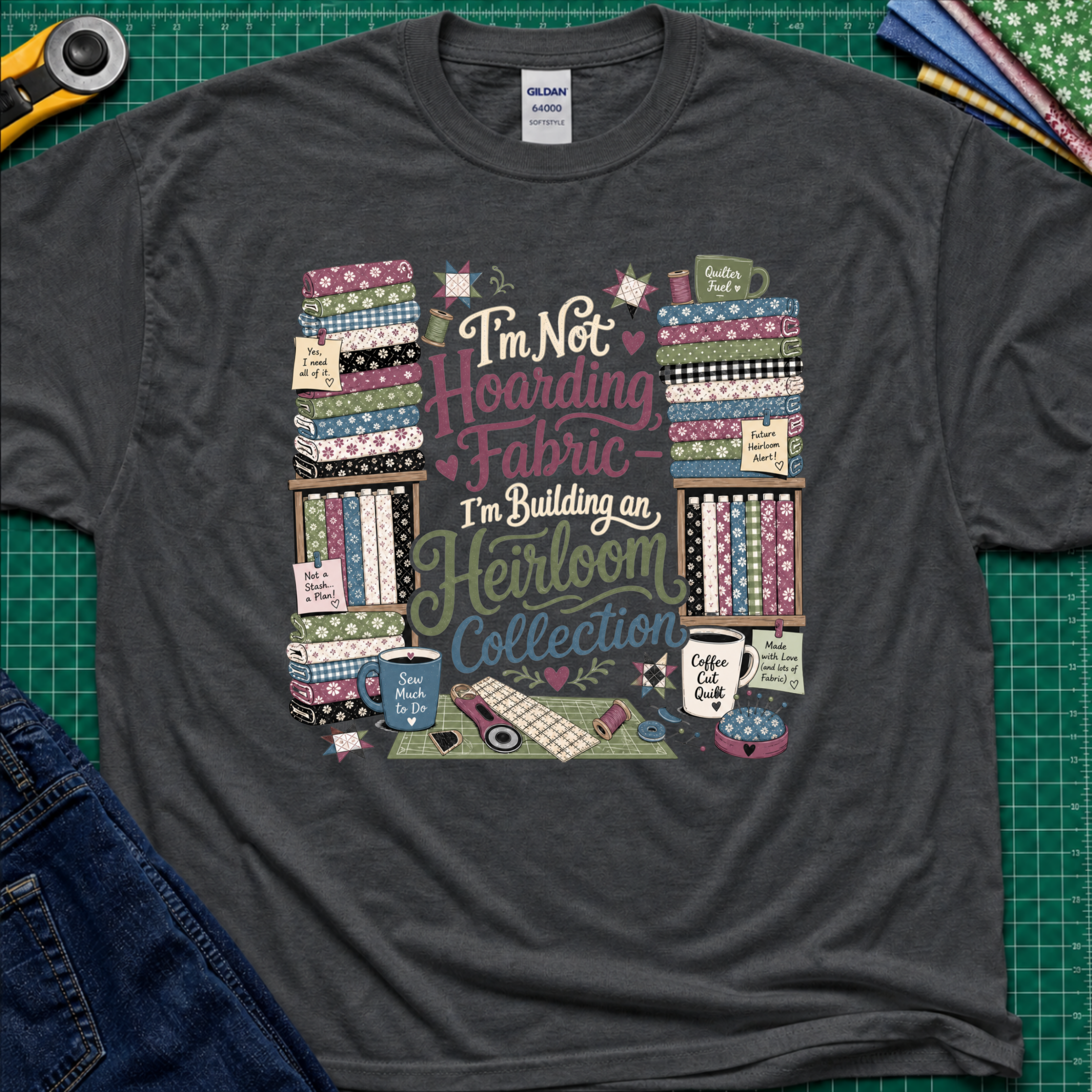 A dark grey Gildan t-shirt  featuring a colorful print with fabric stacks, coffee cups, stars and quilting tools with text that reads, 'I'm Not Hoarding Fabric - I'm Building an Heirloom Collection'. The t-shirt lays flat on a green cutting mat with a rotary cutter, folded fabric and blue jeans partially visible in the corners.
