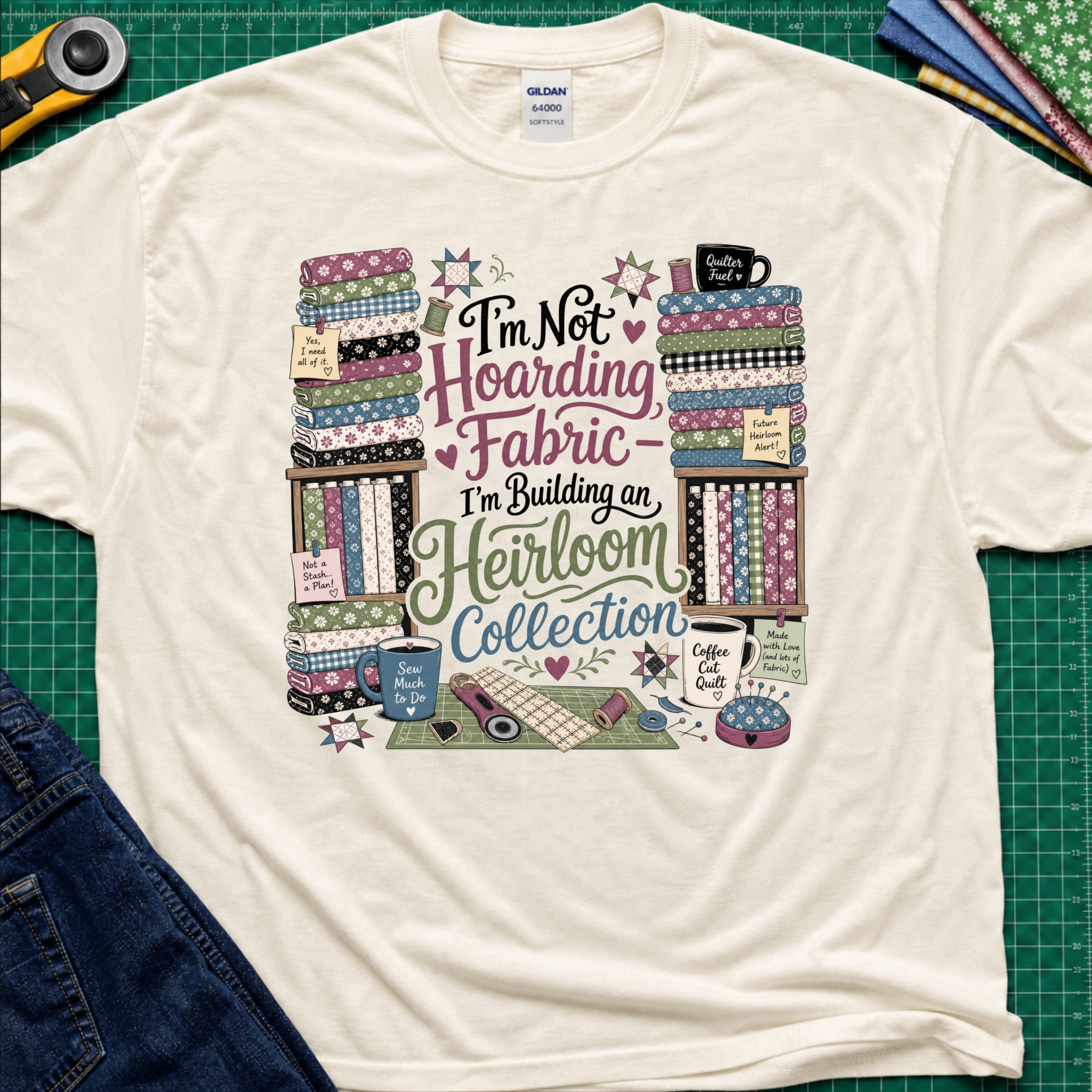 A light cream Gildan t-shirt  featuring a colorful print with fabric stacks, coffee cups, stars and quilting tools with text that reads, 'I'm Not Hoarding Fabric - I'm Building an Heirloom Collection'. The t-shirt lays flat on a green cutting mat with a rotary cutter, folded fabric and blue jeans partially visible in the corners.