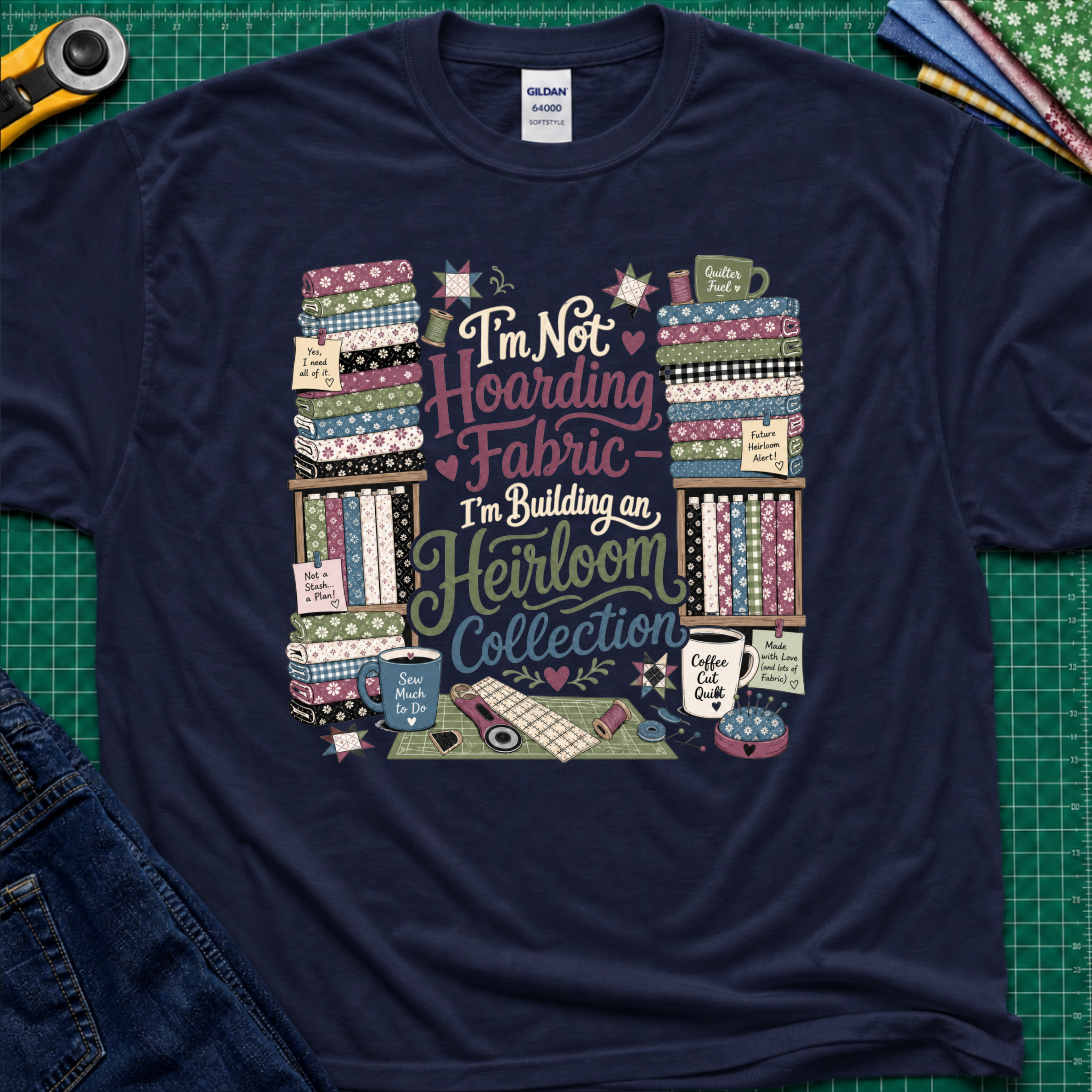 A navy blue Gildan t-shirt  featuring a colorful print with fabric stacks, coffee cups, stars and quilting tools with text that reads, 'I'm Not Hoarding Fabric - I'm Building an Heirloom Collection'. The t-shirt lays flat on a green cutting mat with a rotary cutter, folded fabric and blue jeans partially visible in the corners.