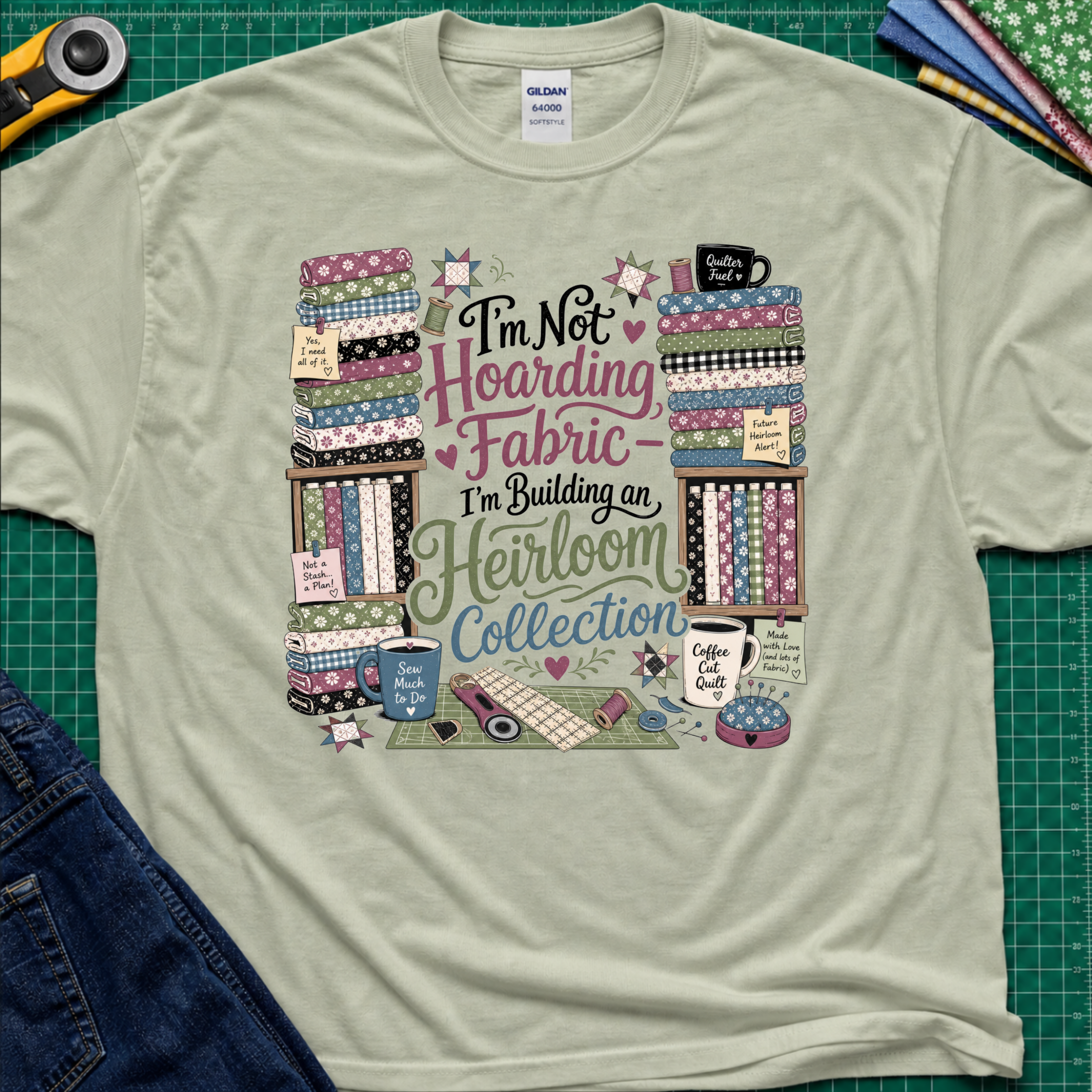 A light sage green Gildan t-shirt  featuring a colorful print with fabric stacks, coffee cups, stars and quilting tools with text that reads, 'I'm Not Hoarding Fabric - I'm Building an Heirloom Collection'. The t-shirt lays flat on a green cutting mat with a rotary cutter, folded fabric and blue jeans partially visible in the corners.