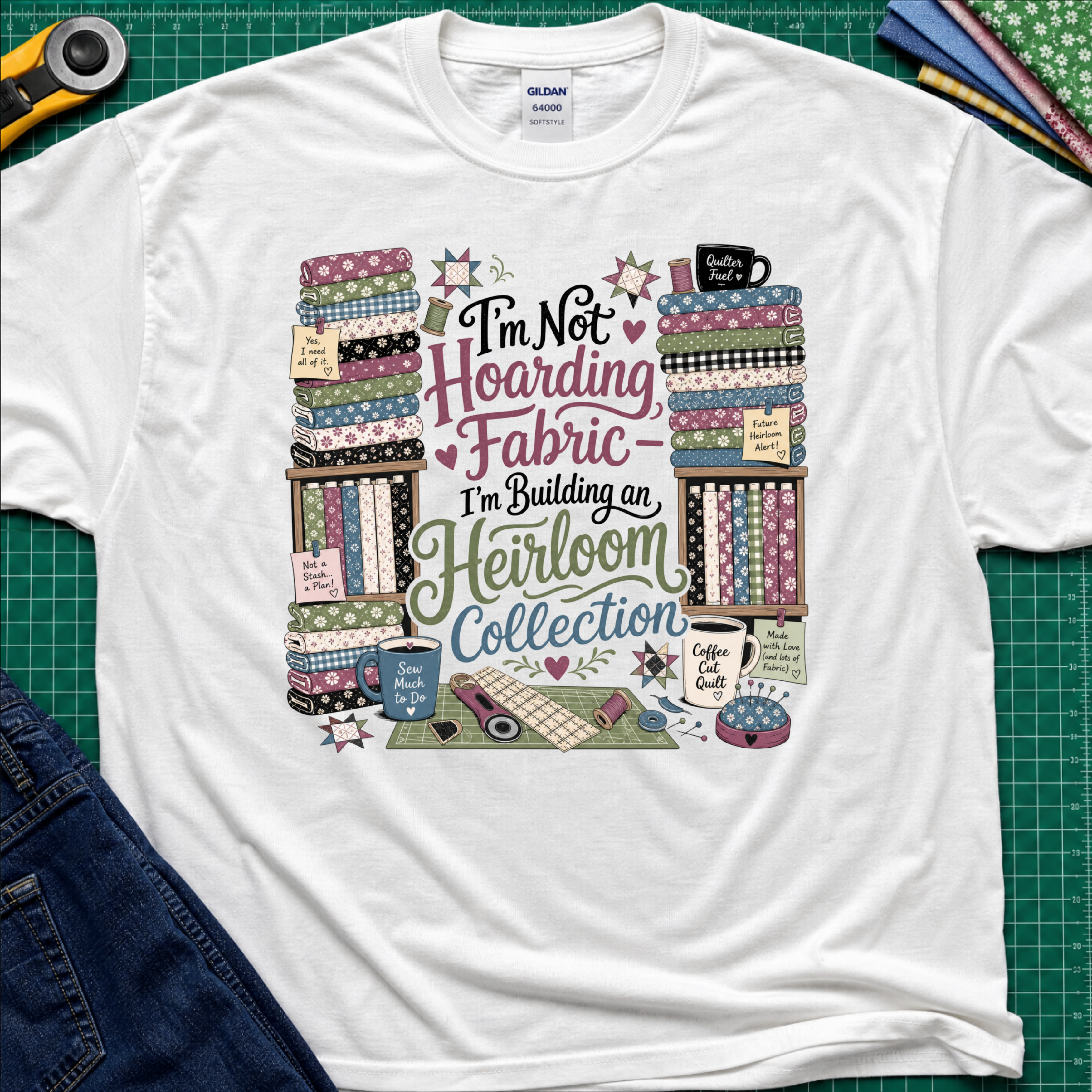 A white Gildan t-shirt  featuring a colorful print with fabric stacks, coffee cups, stars and quilting tools with text that reads, 'I'm Not Hoarding Fabric - I'm Building an Heirloom Collection'. The t-shirt lays flat on a green cutting mat with a rotary cutter, folded fabric and blue jeans partially visible in the corners.