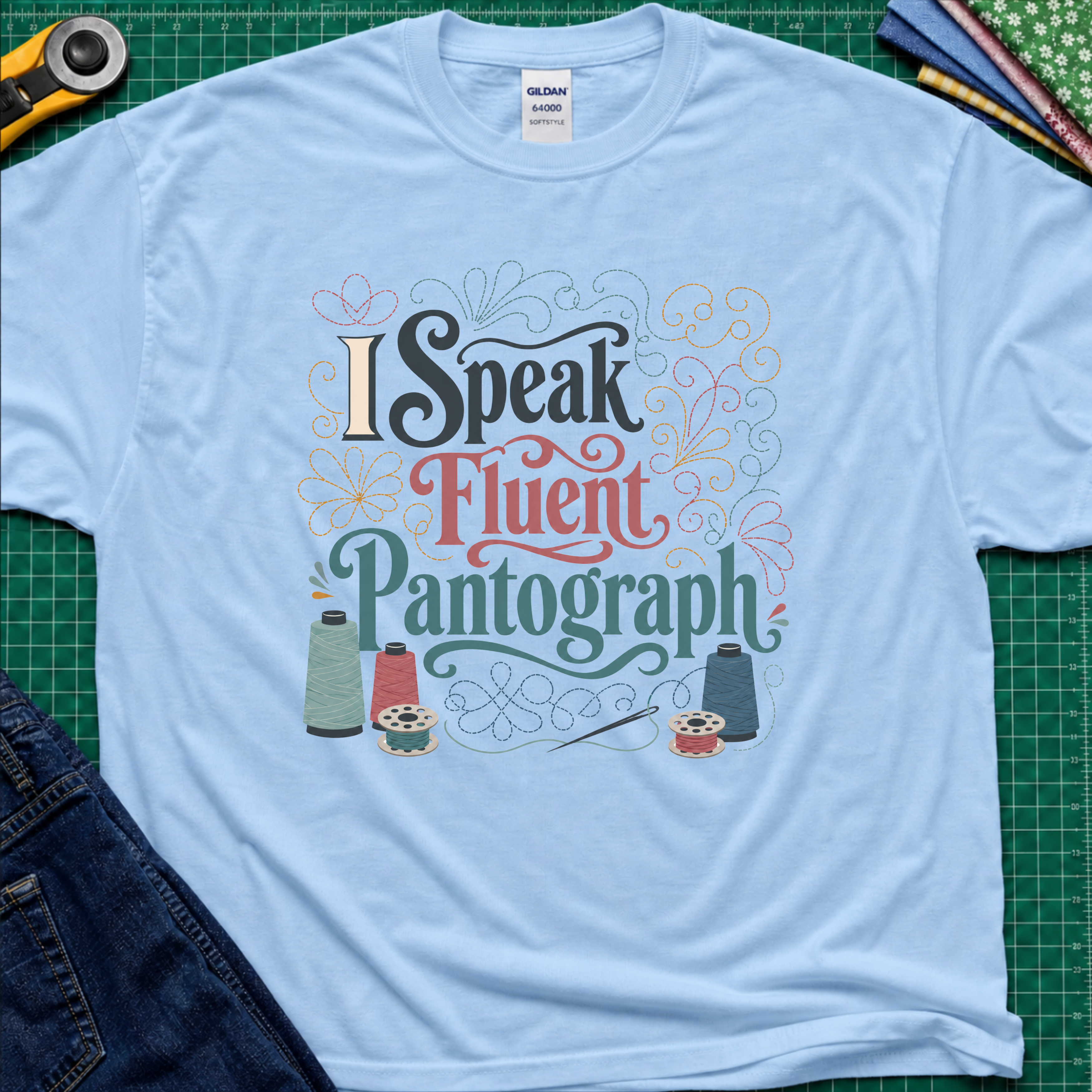 A light blue Gildan t-shirt laid flat on a green cutting mat, featuring a colorful graphic that reads 'I Speak Fluent Pantograph' with decorative swirls and sewing thread spools. A yellow rotary cutter, fabric, and blue jeans are partially visible around the shirt.