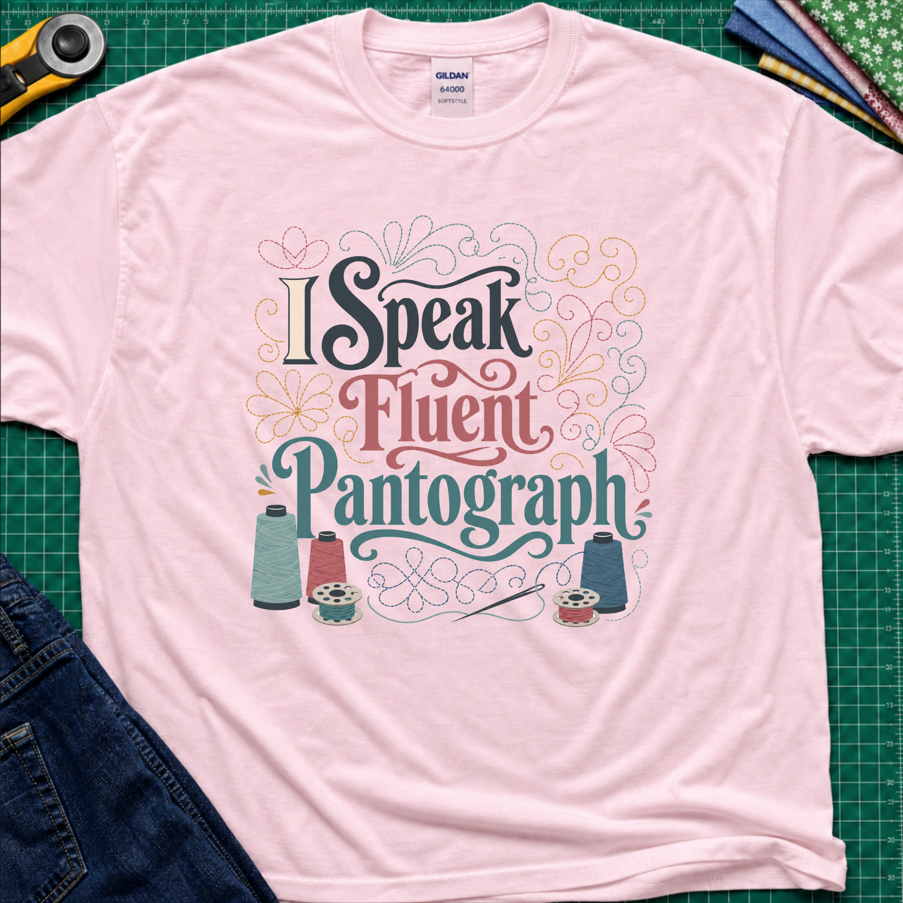 A light pink Gildan t-shirt laid flat on a green cutting mat, featuring a colorful graphic that reads 'I Speak Fluent Pantograph' with decorative swirls and sewing thread spools. A yellow rotary cutter, fabric, and blue jeans are partially visible around the shirt.