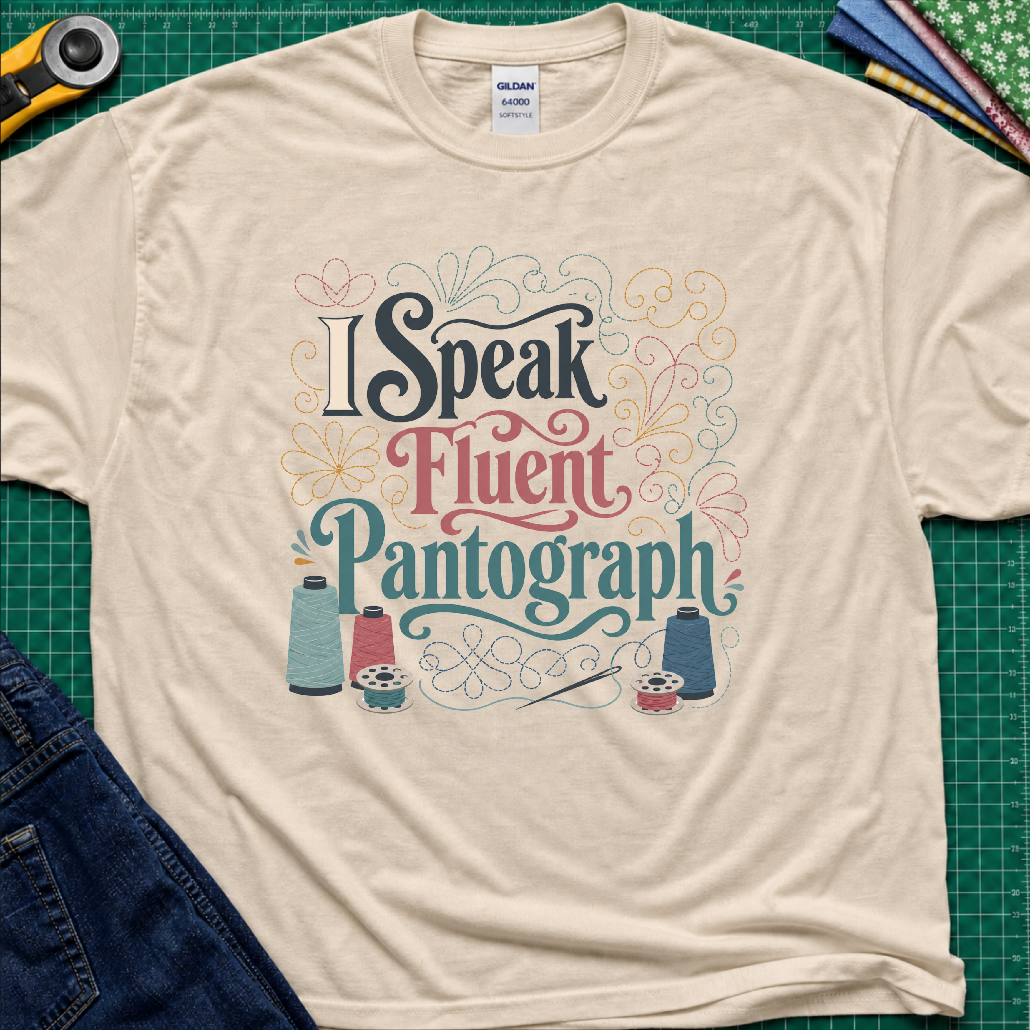 A dark cream Gildan t-shirt laid flat on a green cutting mat, featuring a colorful graphic that reads 'I Speak Fluent Pantograph' with decorative swirls and sewing thread spools. A yellow rotary cutter, fabric, and blue jeans are partially visible around the shirt.