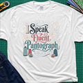 A white Gildan t-shirt laid flat on a green cutting mat, featuring a colorful graphic that reads 'I Speak Fluent Pantograph' with decorative swirls and sewing thread spools. A yellow rotary cutter, fabric, and blue jeans are partially visible around the shirt.