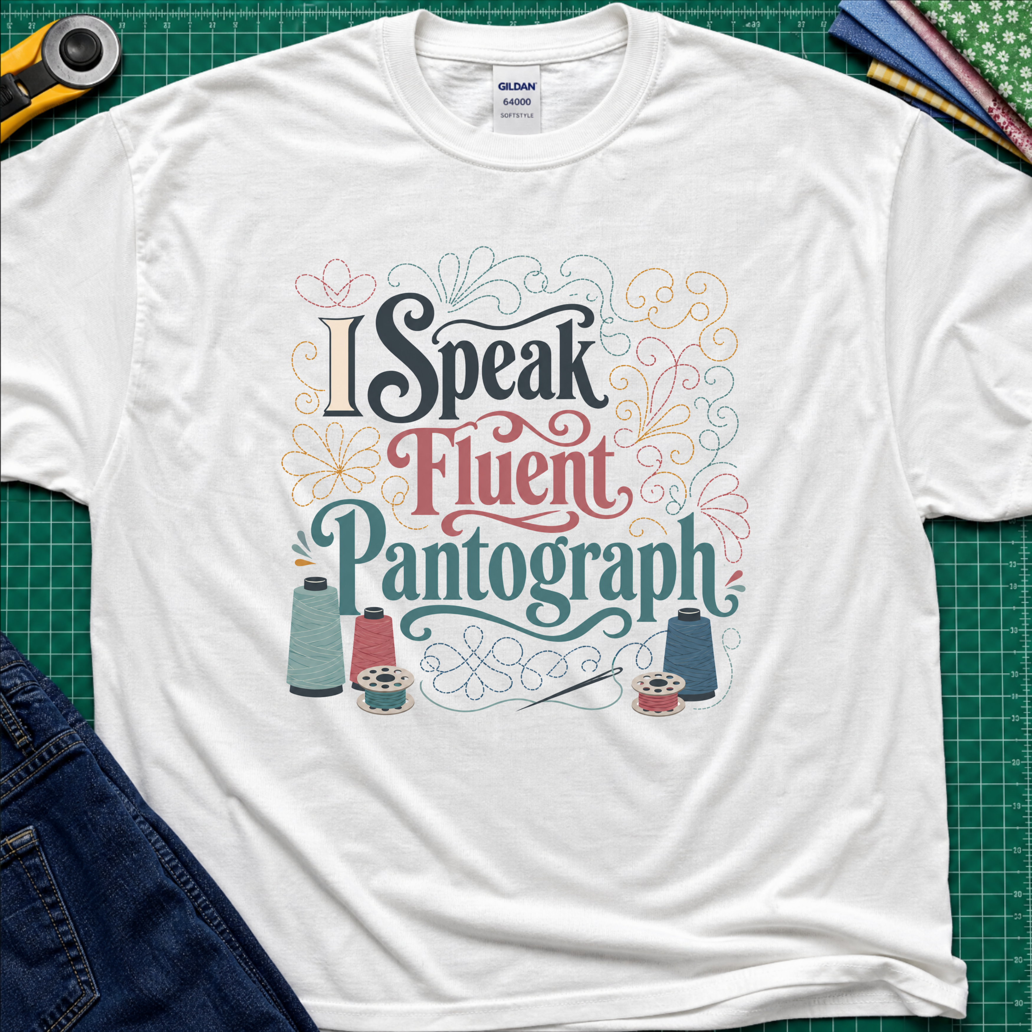 A white Gildan t-shirt laid flat on a green cutting mat, featuring a colorful graphic that reads 'I Speak Fluent Pantograph' with decorative swirls and sewing thread spools. A yellow rotary cutter, fabric, and blue jeans are partially visible around the shirt.