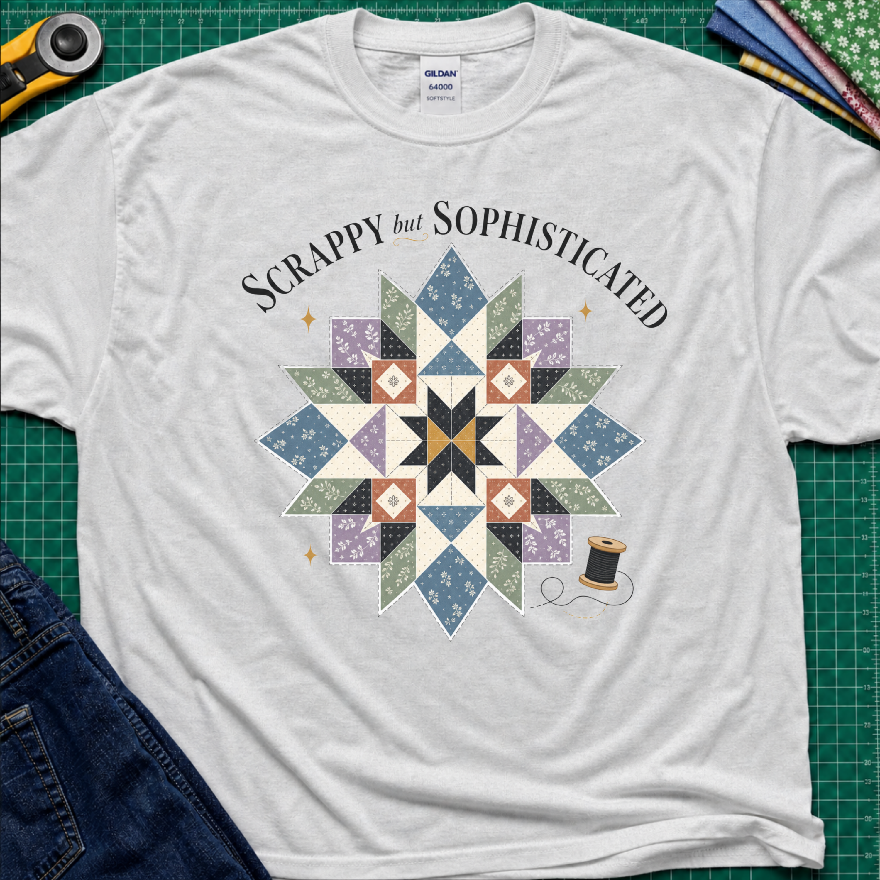 An ice grey Gildan t-shirt with a colorful quilted starburst graphic and the text 'Scrappy but Sophisticated' printed on the front, styled on a green cutting mat with sewing tools and denim.