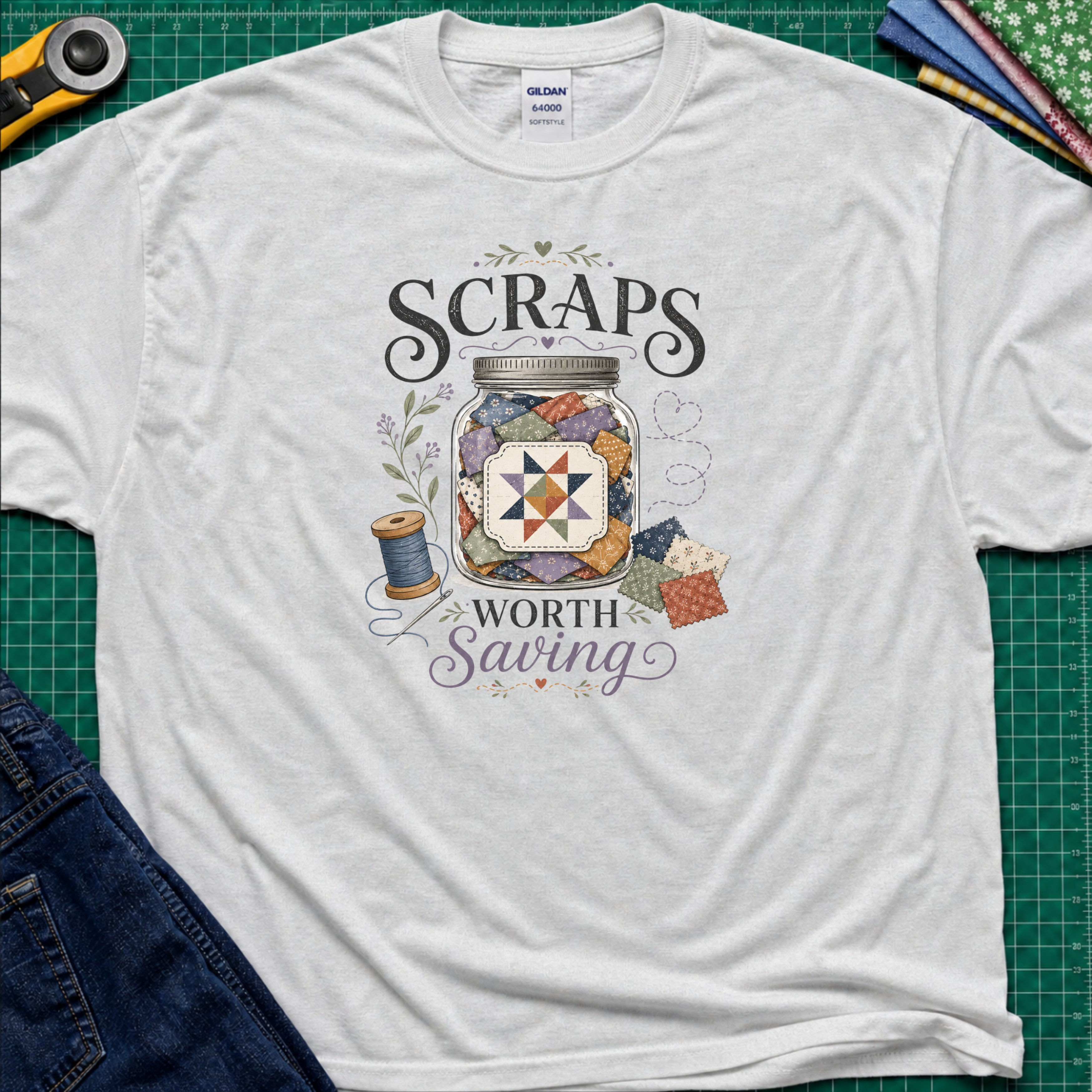 A ice grey Gildan t-shirt laid flat on a green cutting mat, featuring a graphic design that reads 'SCRAPS WORTH Saving' around an illustration of a jar filled with scraps, thread spool, and fabric pieces. A rotary cutter, scissors, and folded fabrics are visible around the shirt as styling props.