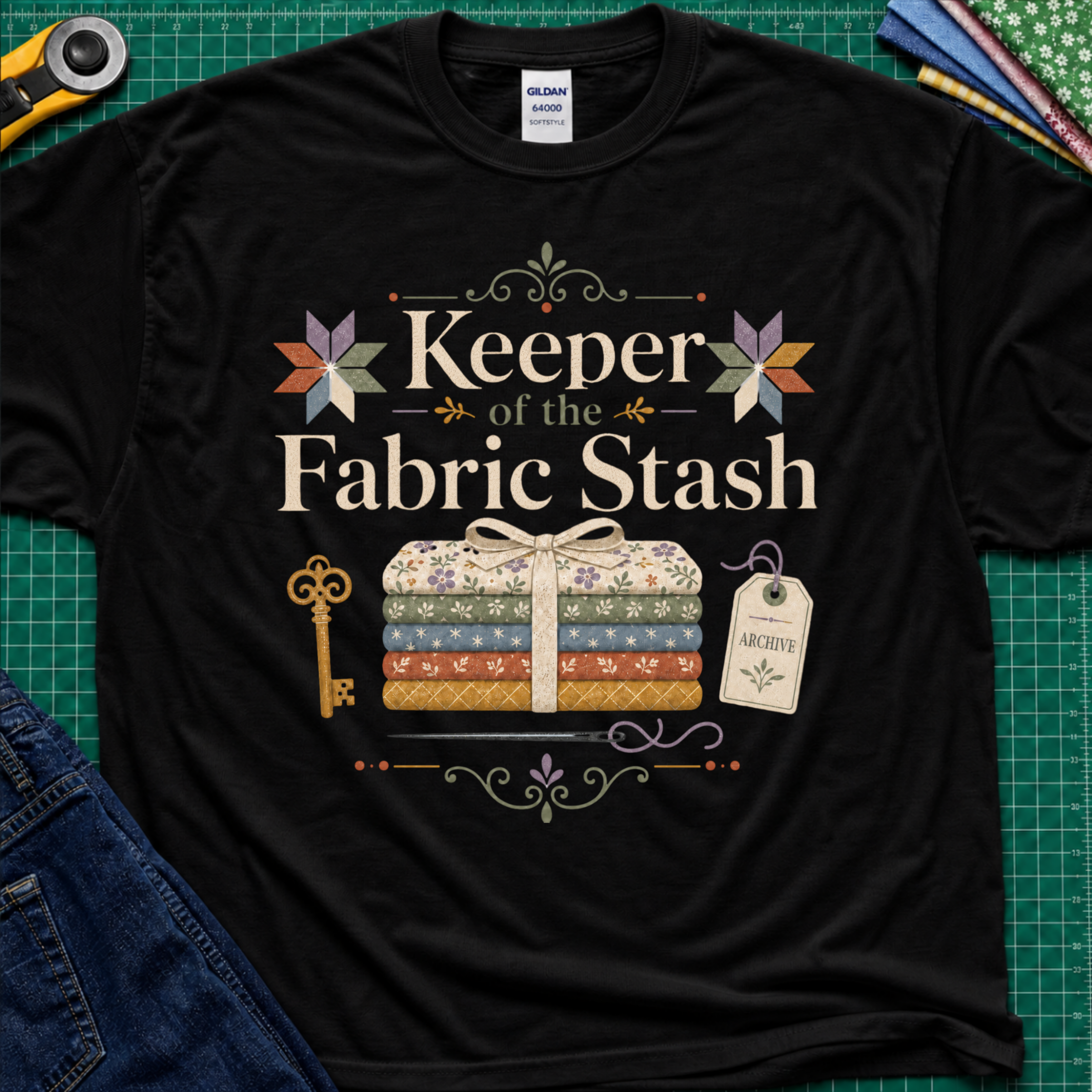 A black Gildan t-shirt featuring the words 'Keeper of the Fabric Stash' with a fabric stack tied in ribbon, patchwork stars, a key, and archive tag. Shirt lays flat on a green cutting mat with rotary cutter, folded fat quarters, and denim jeans partially visible in the corners.
