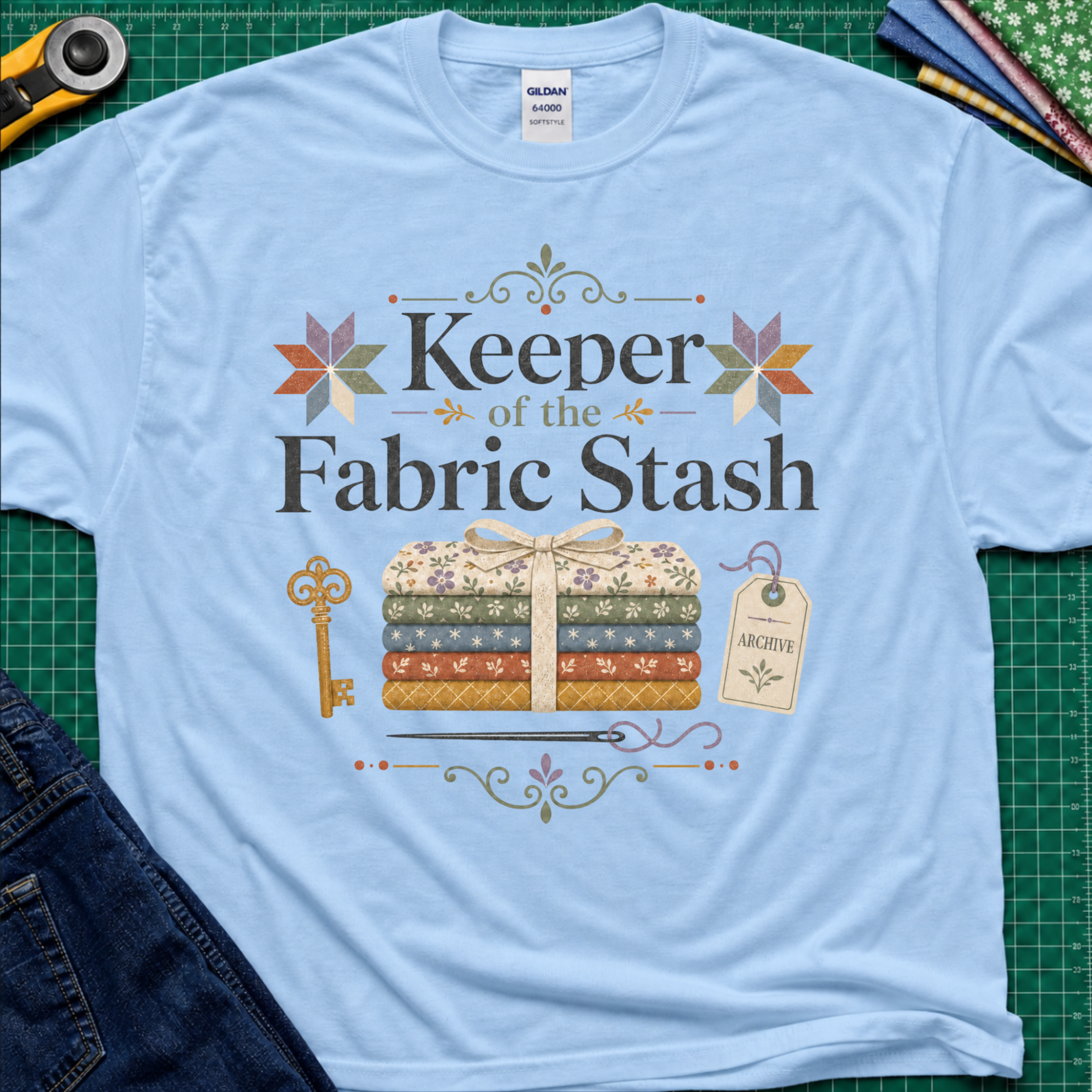 A light blue Gildan t-shirt featuring the words 'Keeper of the Fabric Stash' with a fabric stack tied in ribbon, patchwork stars, a key, and archive tag. Shirt lays flat on a green cutting mat with rotary cutter, folded fat quarters, and denim jeans partially visible in the corners.