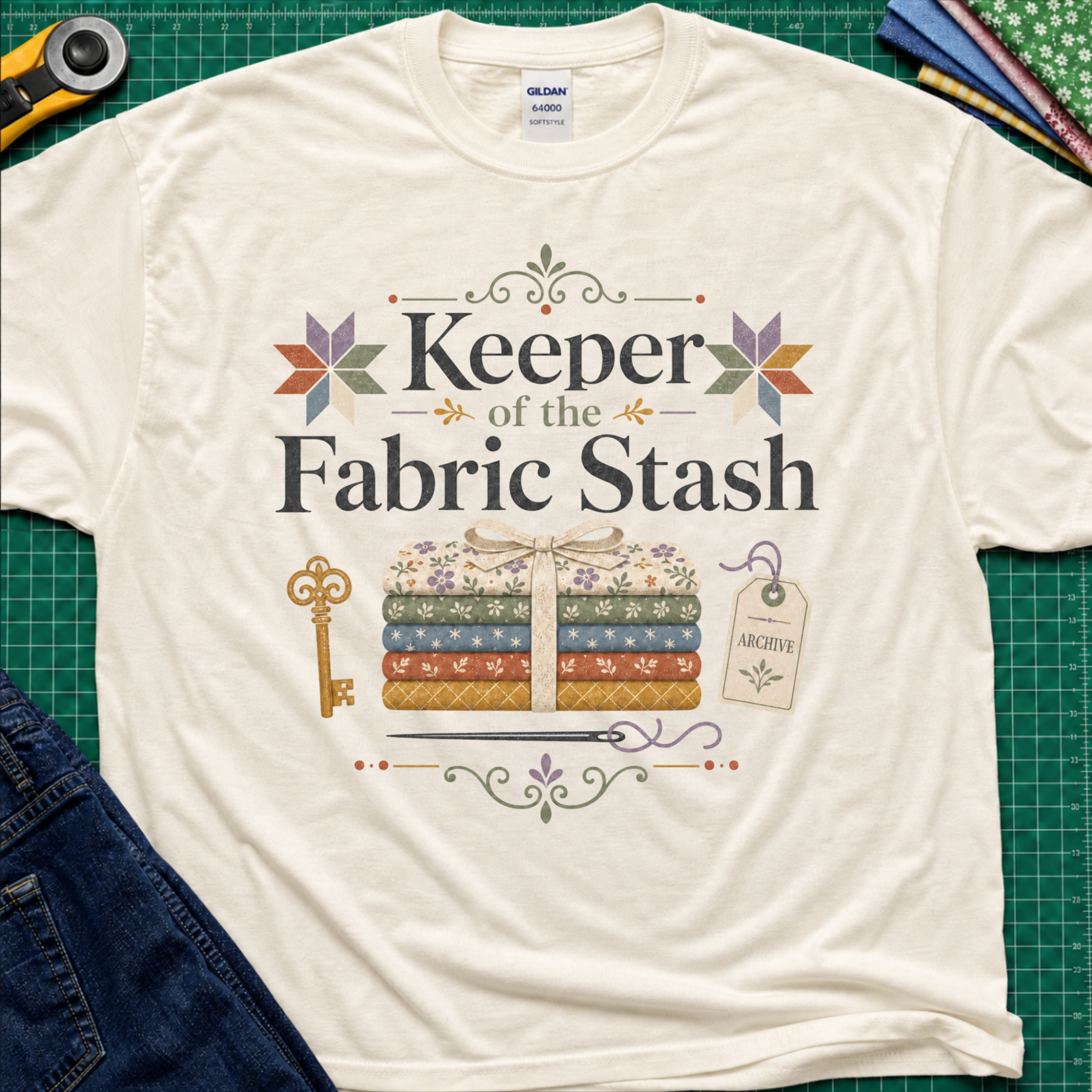 A light cream Gildan t-shirt featuring the words 'Keeper of the Fabric Stash' with a fabric stack tied in ribbon, patchwork stars, a key, and archive tag. Shirt lays flat on a green cutting mat with rotary cutter, folded fat quarters, and denim jeans partially visible in the corners.