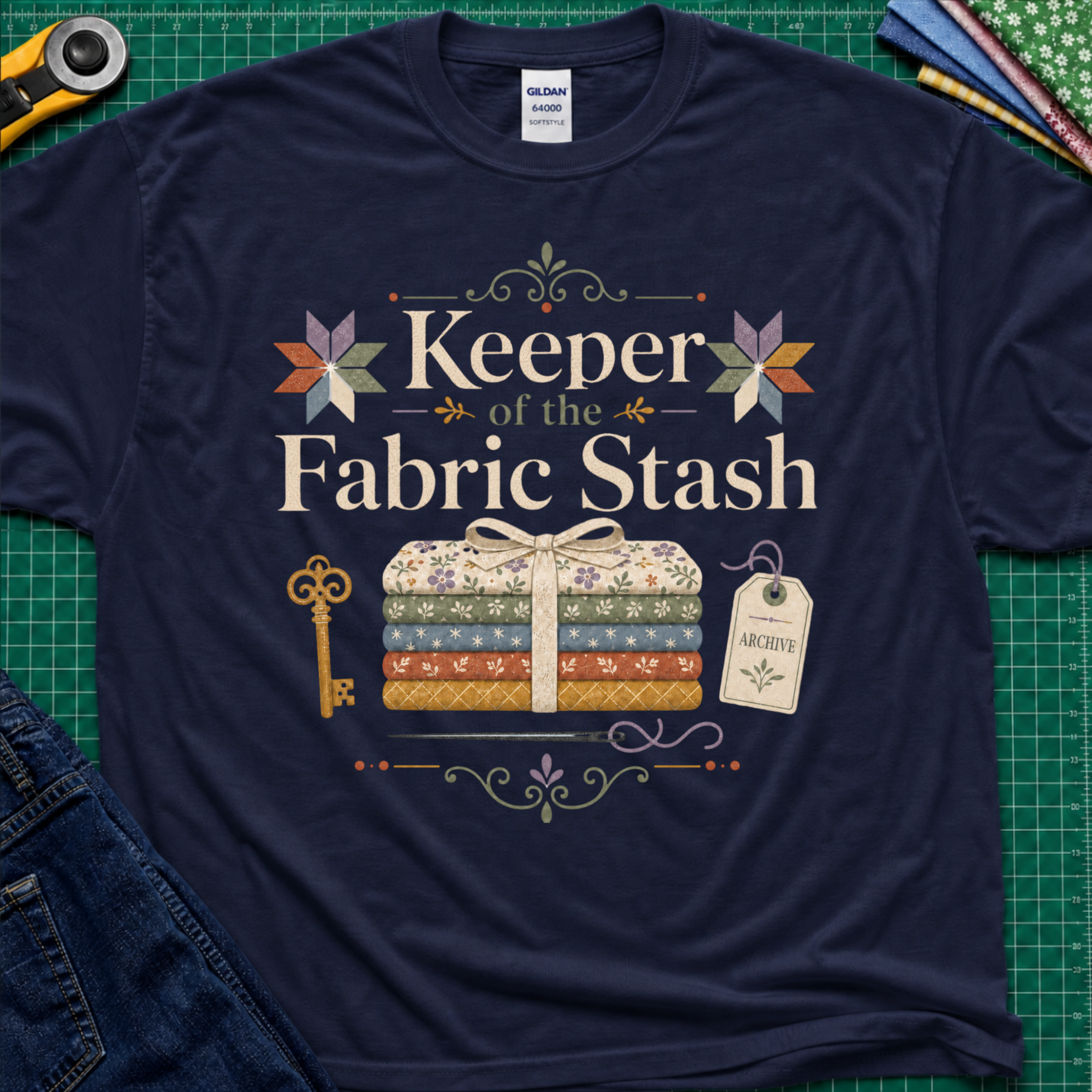 A navy blue Gildan t-shirt featuring the words 'Keeper of the Fabric Stash' with a fabric stack tied in ribbon, patchwork stars, a key, and archive tag. Shirt lays flat on a green cutting mat with rotary cutter, folded fat quarters, and denim jeans partially visible in the corners.