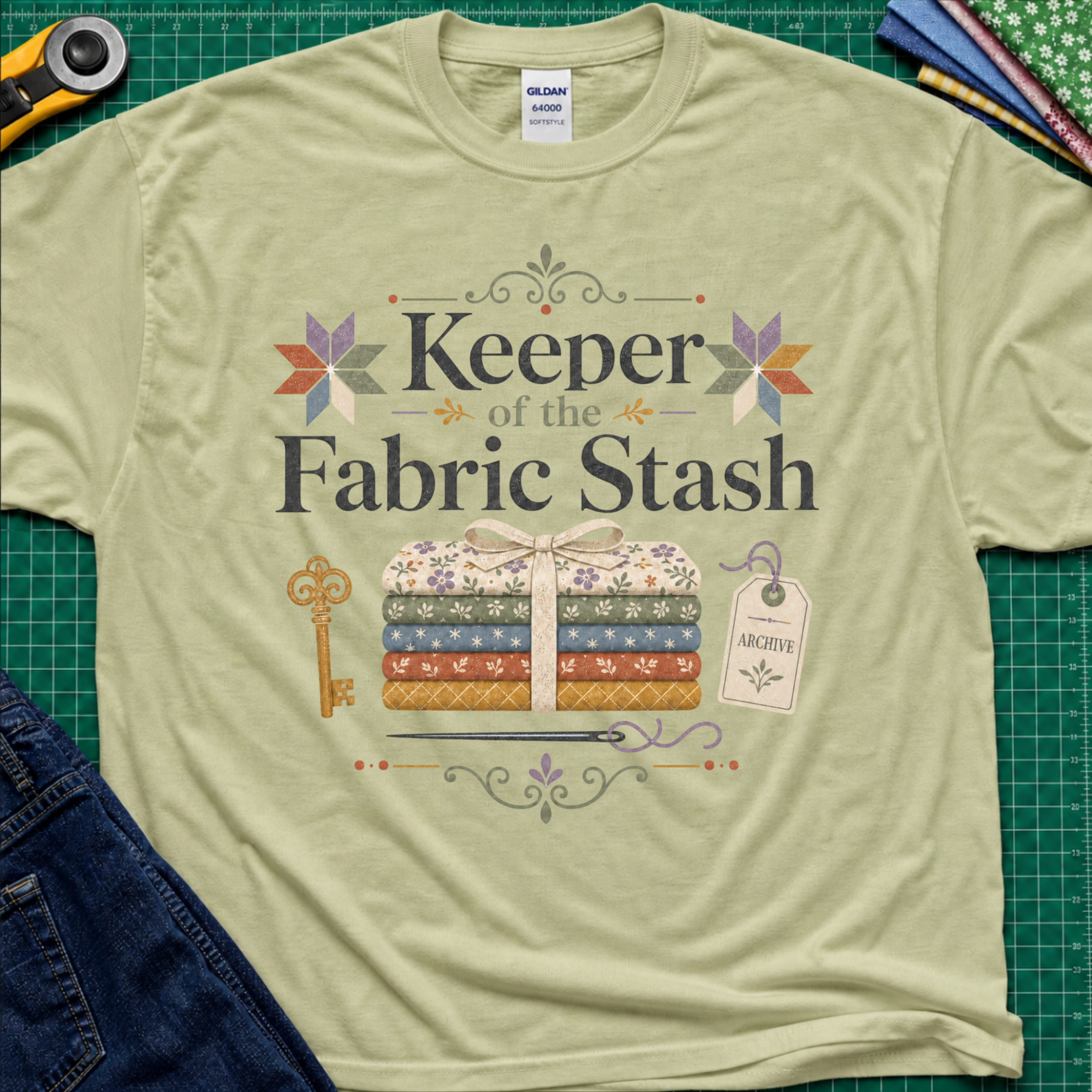 A light pistachio green Gildan t-shirt featuring the words 'Keeper of the Fabric Stash' with a fabric stack tied in ribbon, patchwork stars, a key, and archive tag. Shirt lays flat on a green cutting mat with rotary cutter, folded fat quarters, and denim jeans partially visible in the corners.