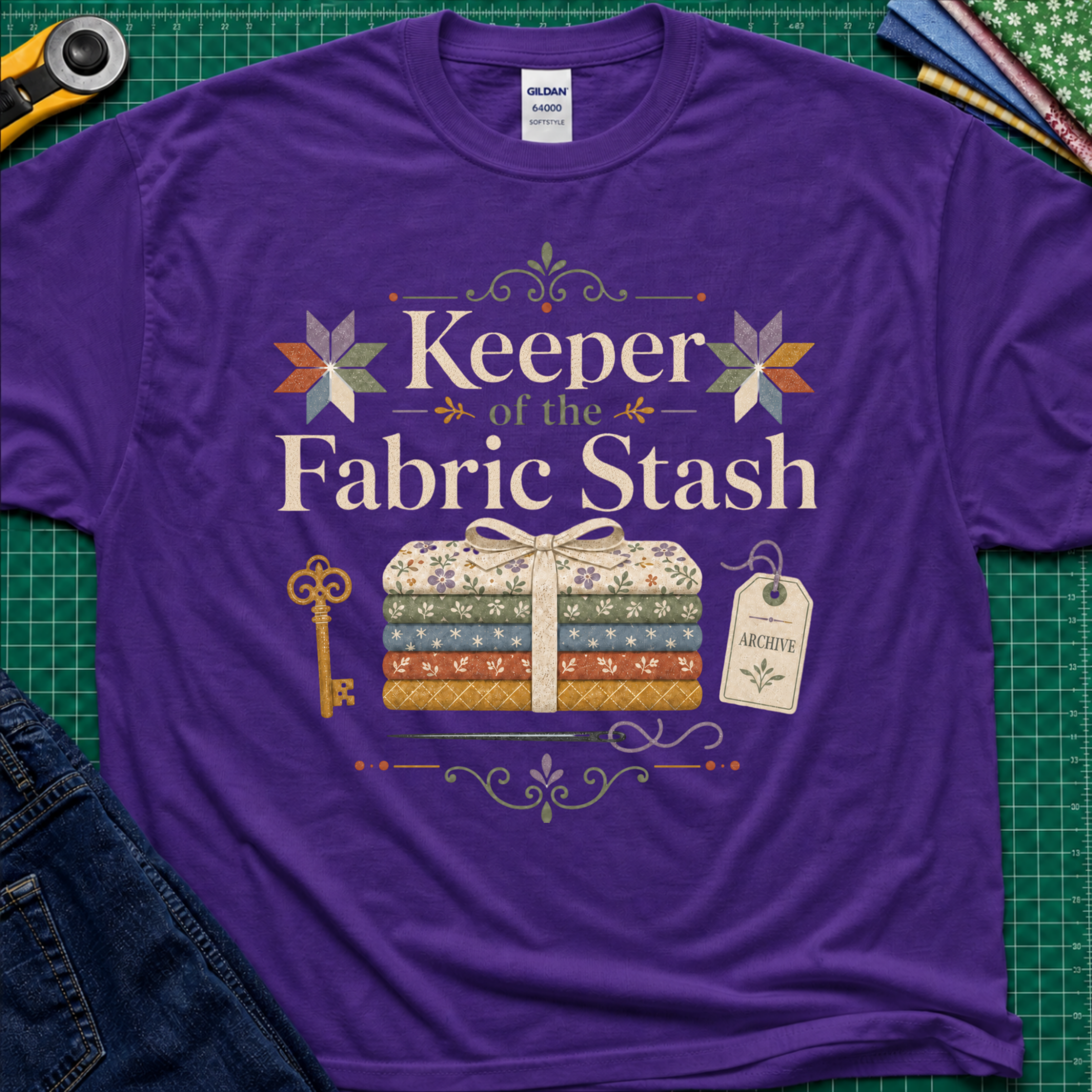 A purple Gildan t-shirt featuring the words 'Keeper of the Fabric Stash' with a fabric stack tied in ribbon, patchwork stars, a key, and archive tag. Shirt lays flat on a green cutting mat with rotary cutter, folded fat quarters, and denim jeans partially visible in the corners.