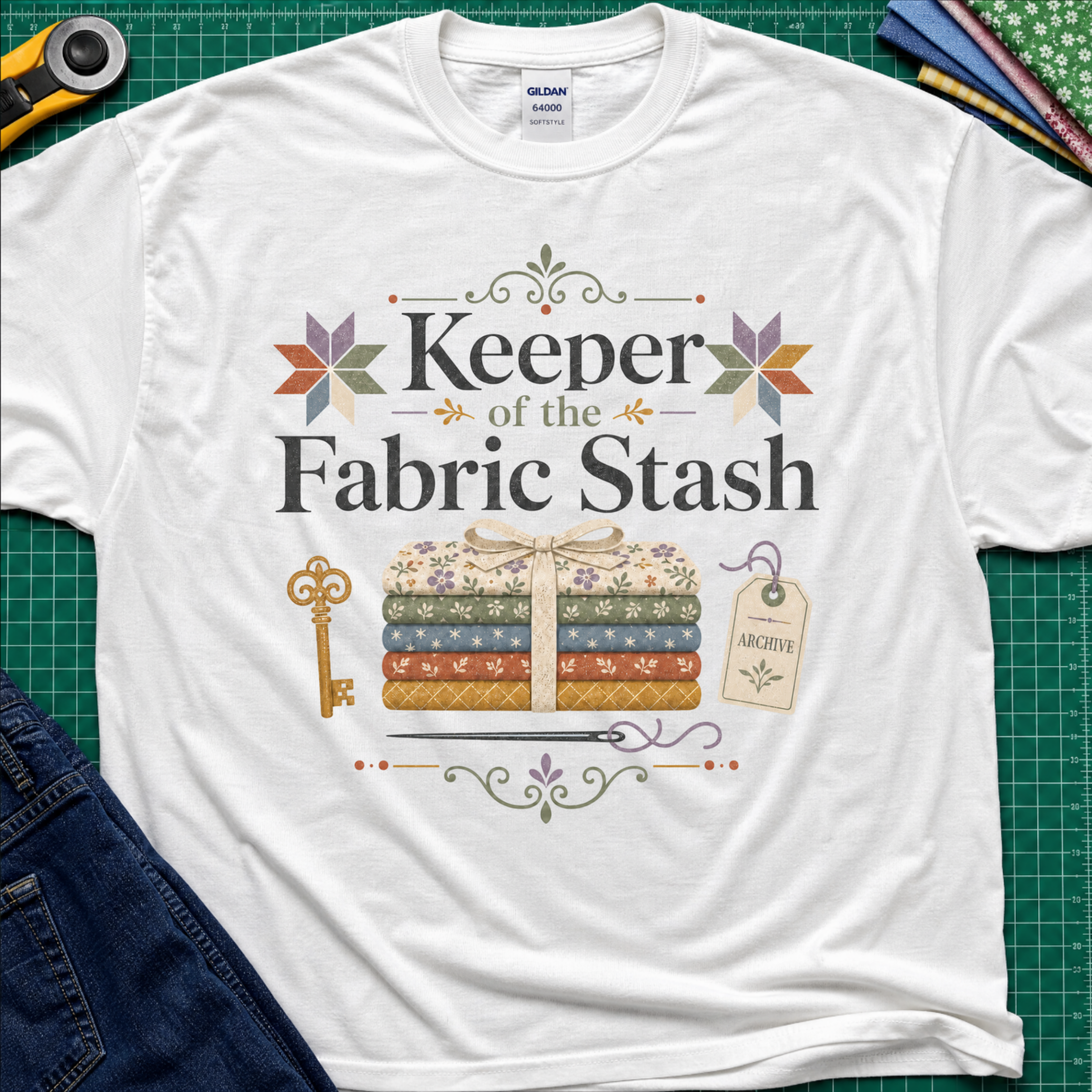 A white Gildan t-shirt featuring the words 'Keeper of the Fabric Stash' with a fabric stack tied in ribbon, patchwork stars, a key, and archive tag. Shirt lays flat on a green cutting mat with rotary cutter, folded fat quarters, and denim jeans partially visible in the corners.