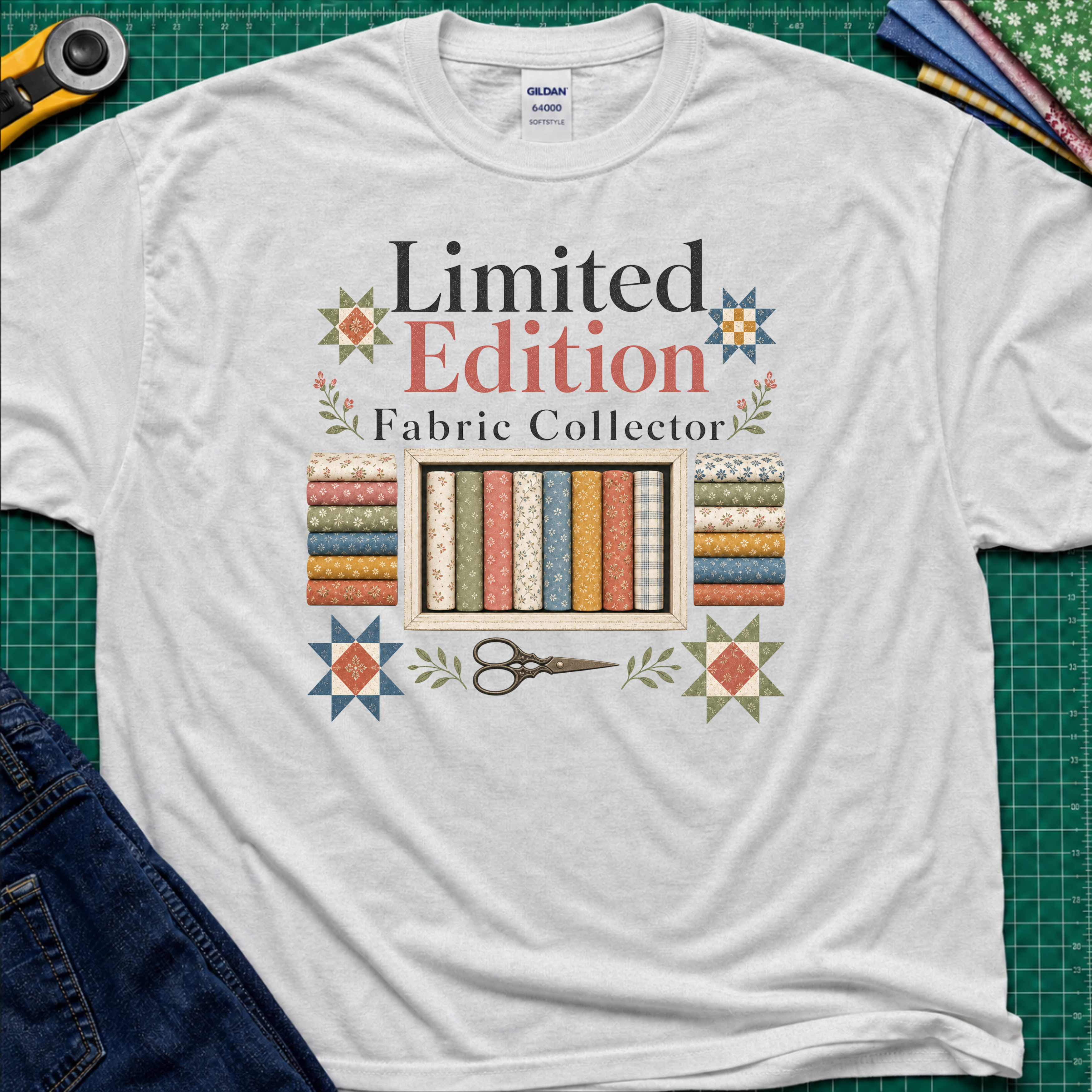 A light grey Gildan t-shirt featuring a colorful graphic that reads 'Limited Edition Fabric Collector' with fabric rolls, scissors, and decorative stars. The shirt is laying on a green cutting mat with quilting supplies and blue jeans around the outside.