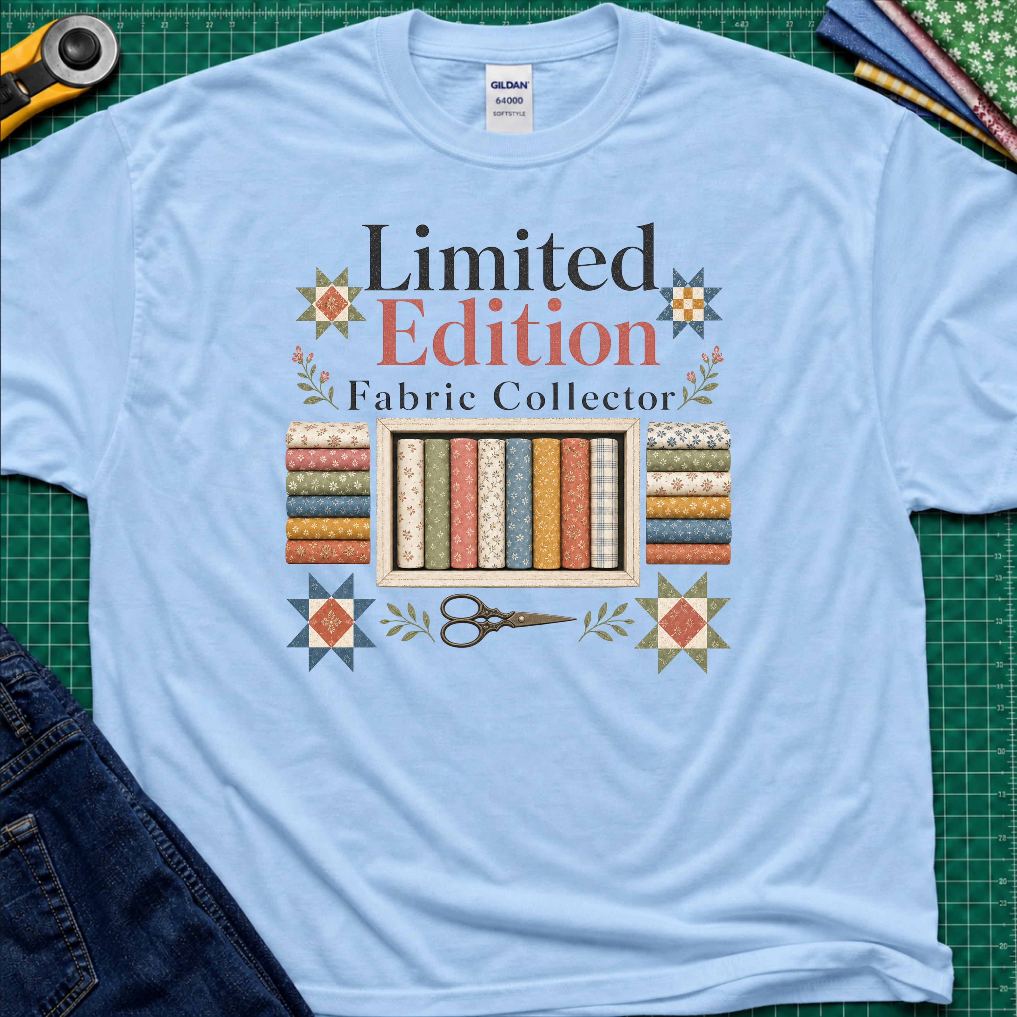A light blue Gildan t-shirt featuring a colorful graphic that reads 'Limited Edition Fabric Collector' with fabric rolls, scissors, and decorative stars. The shirt is laying on a green cutting mat with quilting supplies and blue jeans around the outside.