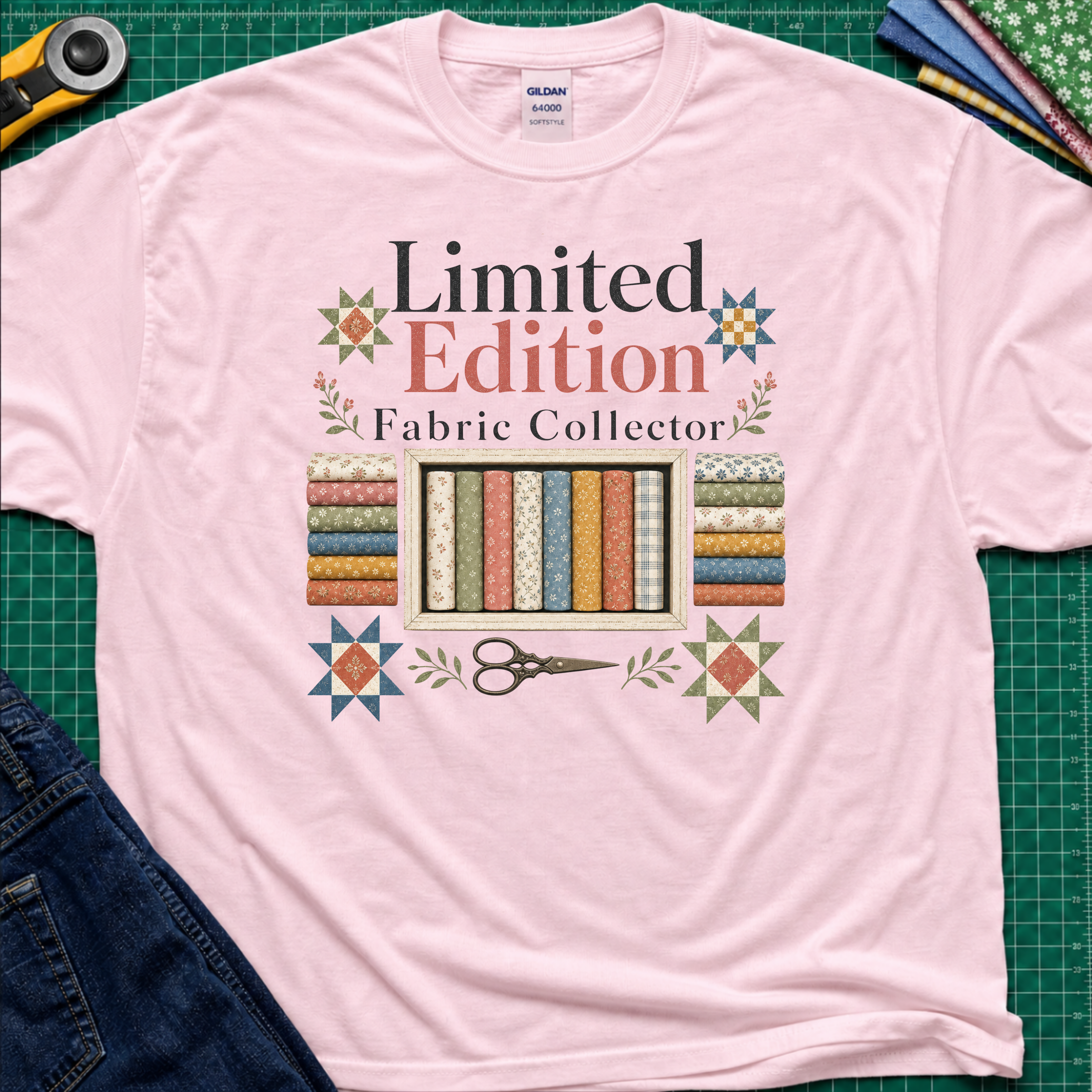 A light pink Gildan t-shirt featuring a colorful graphic that reads 'Limited Edition Fabric Collector' with fabric rolls, scissors, and decorative stars. The shirt is laying on a green cutting mat with quilting supplies and blue jeans around the outside.