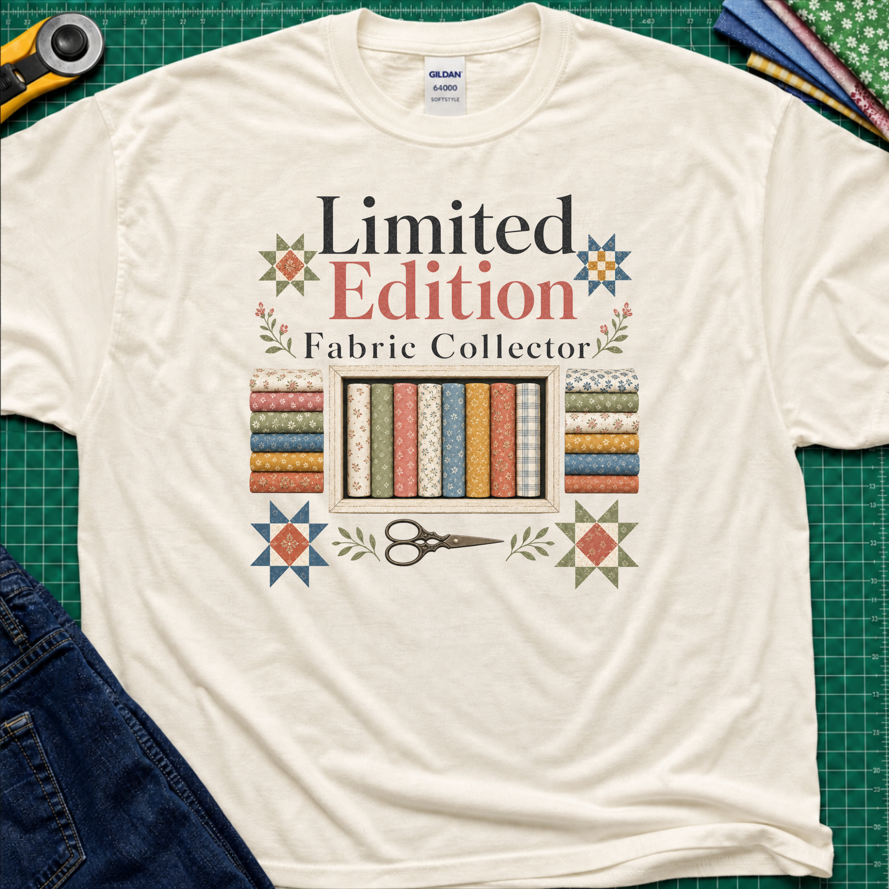 A light cream Gildan t-shirt featuring a colorful graphic that reads 'Limited Edition Fabric Collector' with fabric rolls, scissors, and decorative stars. The shirt is laying on a green cutting mat with quilting supplies and blue jeans around the outside.